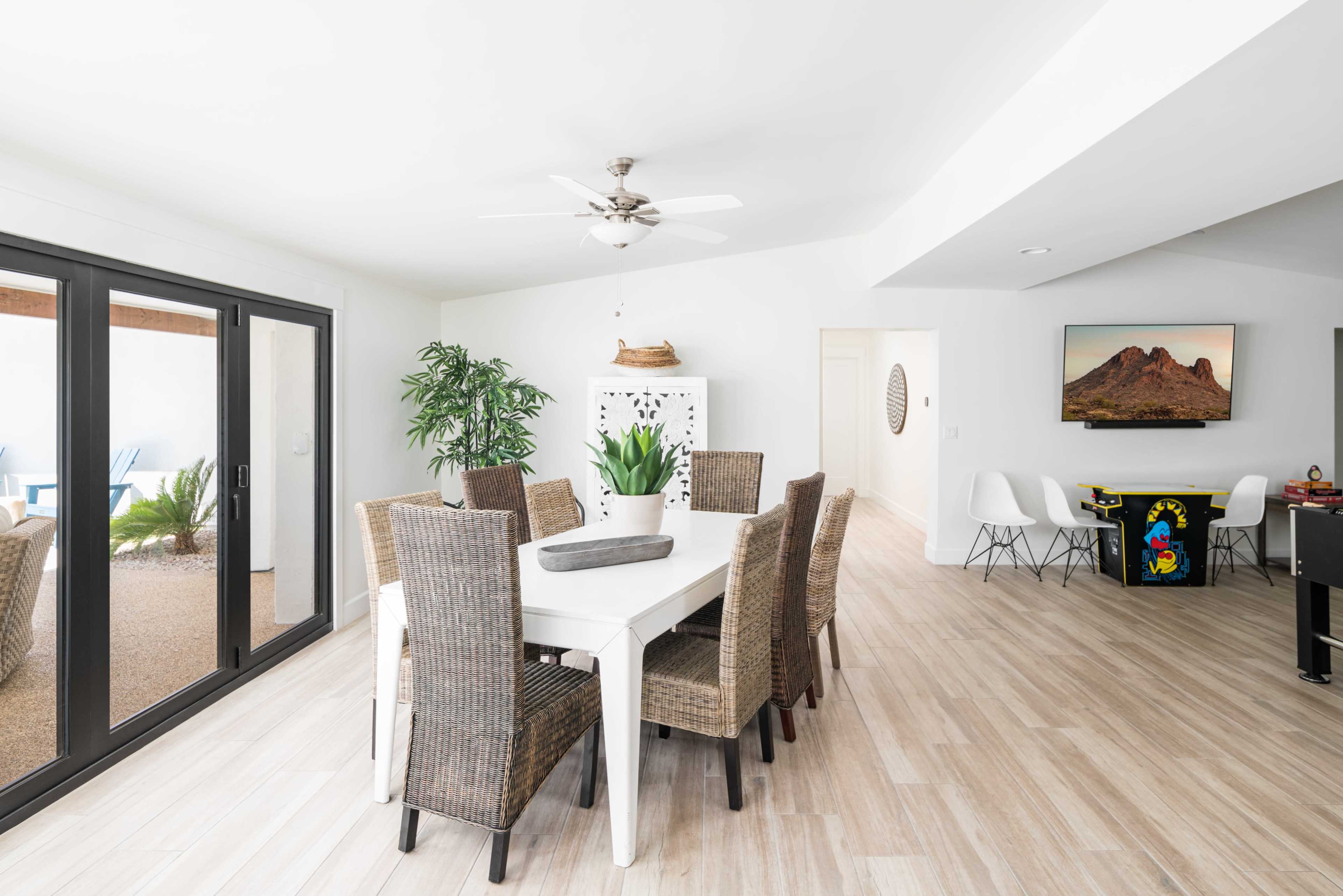 A modern dining area features a large white table surrounded by woven chairs, with a view of a patio through glass sliding doors and a kitchen visible in the background.