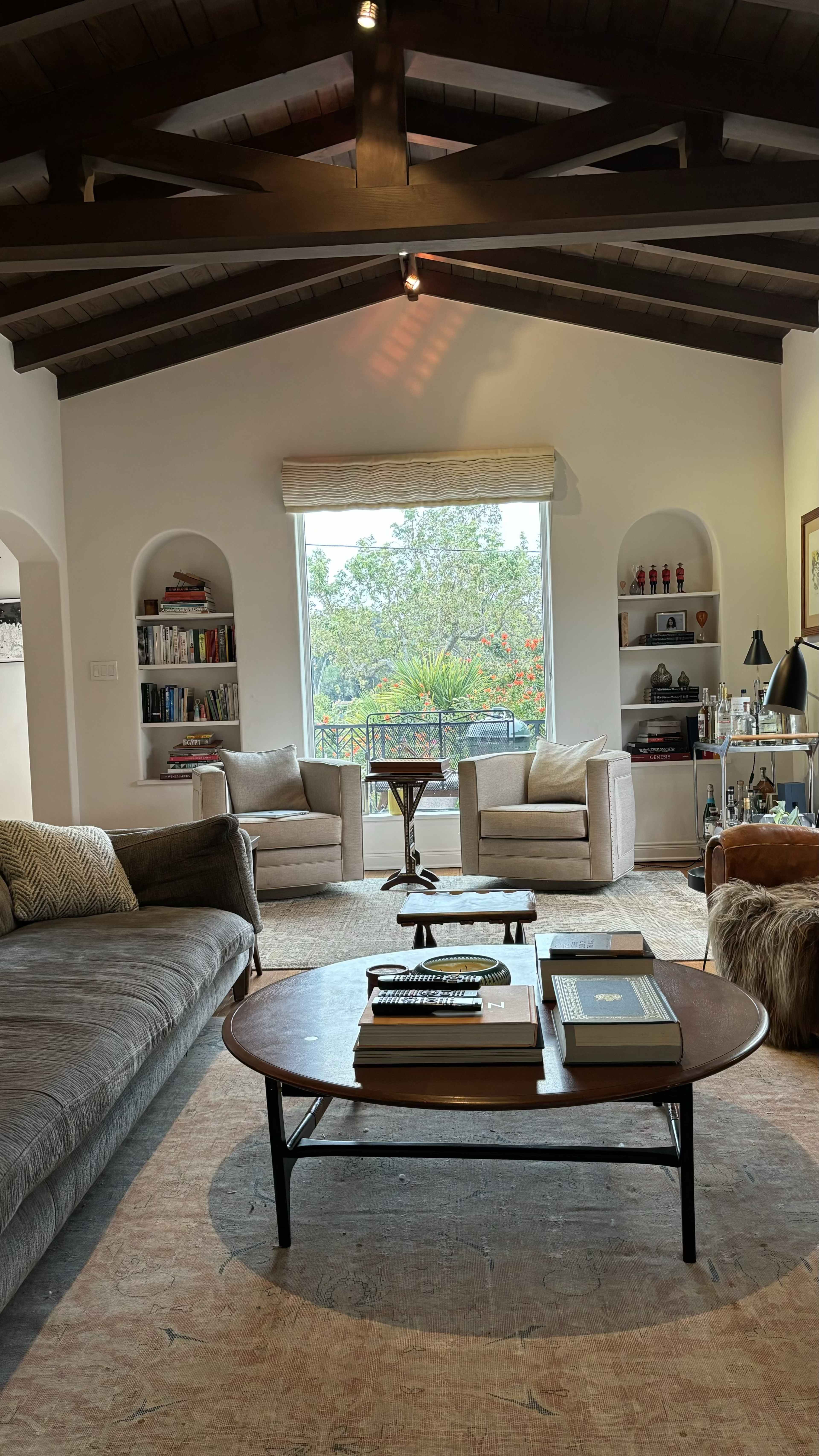 The image depicts a cozy living room featuring two armchairs, a coffee table with books, and a large window providing natural light, surrounded by bookshelves and decorative items.