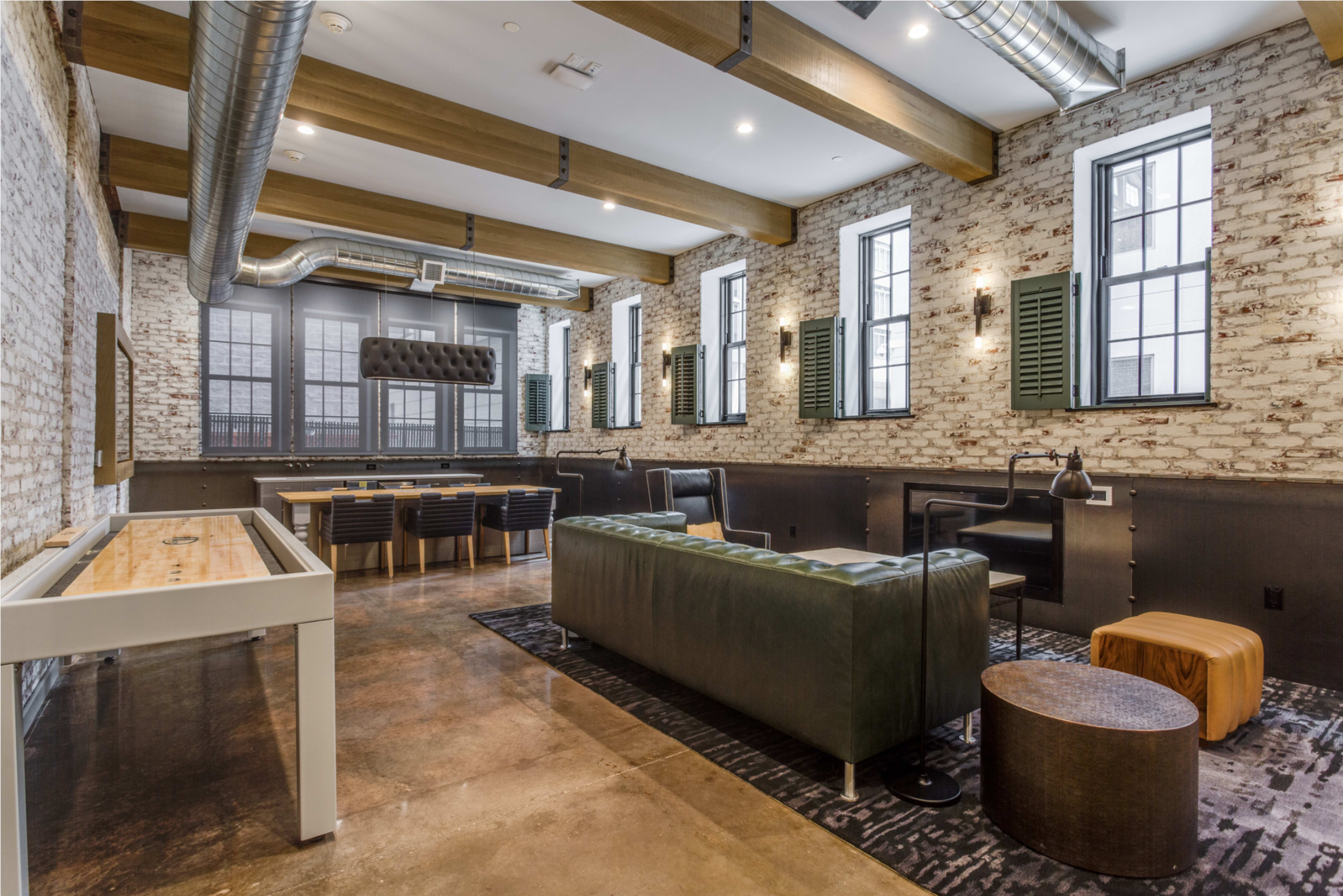 The image shows a modern loft-style room with exposed brick walls, large windows, and a mix of seating options, including a green leather couch and a wooden table.