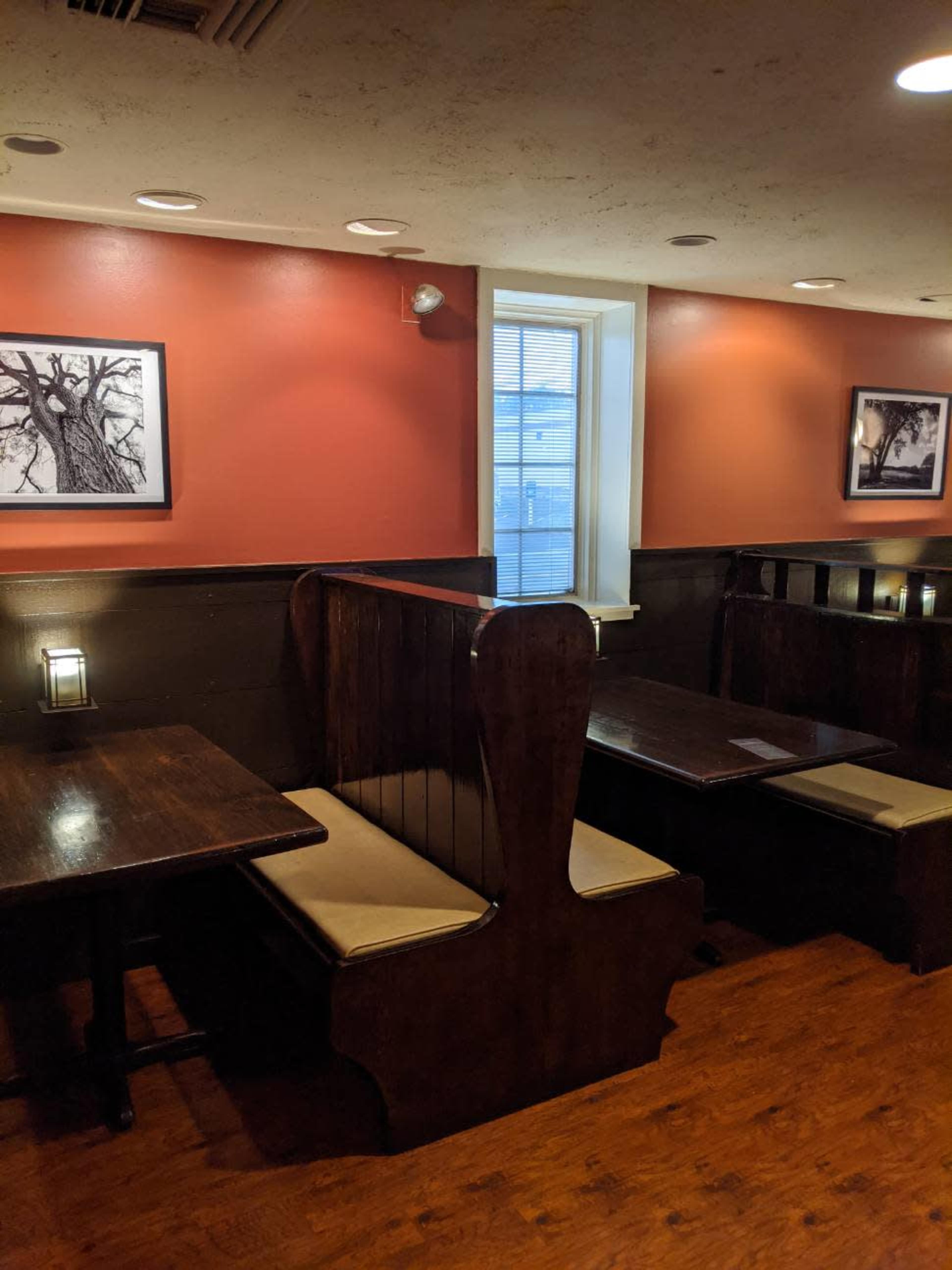 A restaurant interior with dark wooden tables, a window, and booths along the walls, featuring framed black and white photographs.