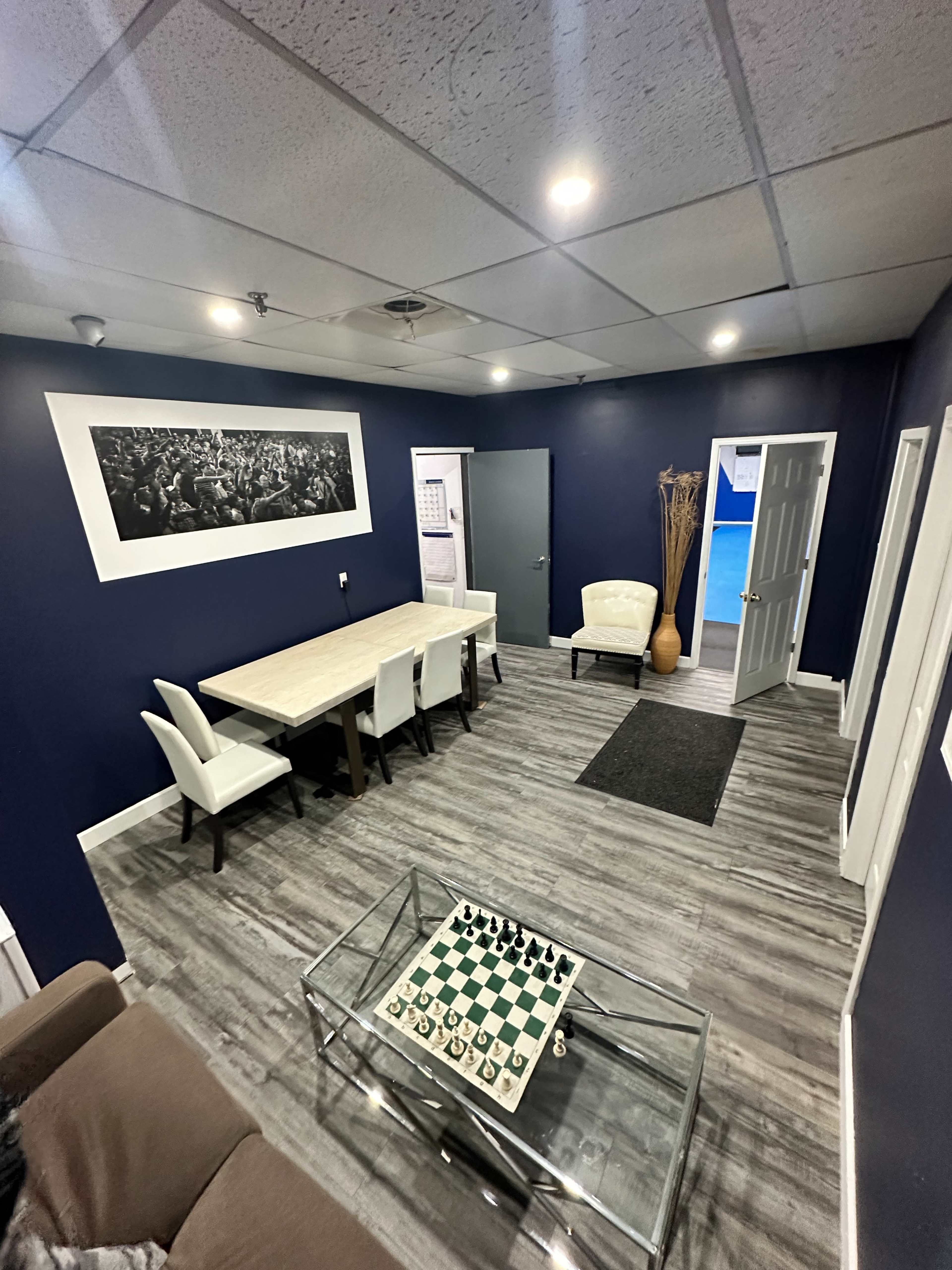 The image shows a modern office or meeting space with a dining table and chairs, a chessboard on a glass coffee table, and navy blue walls.