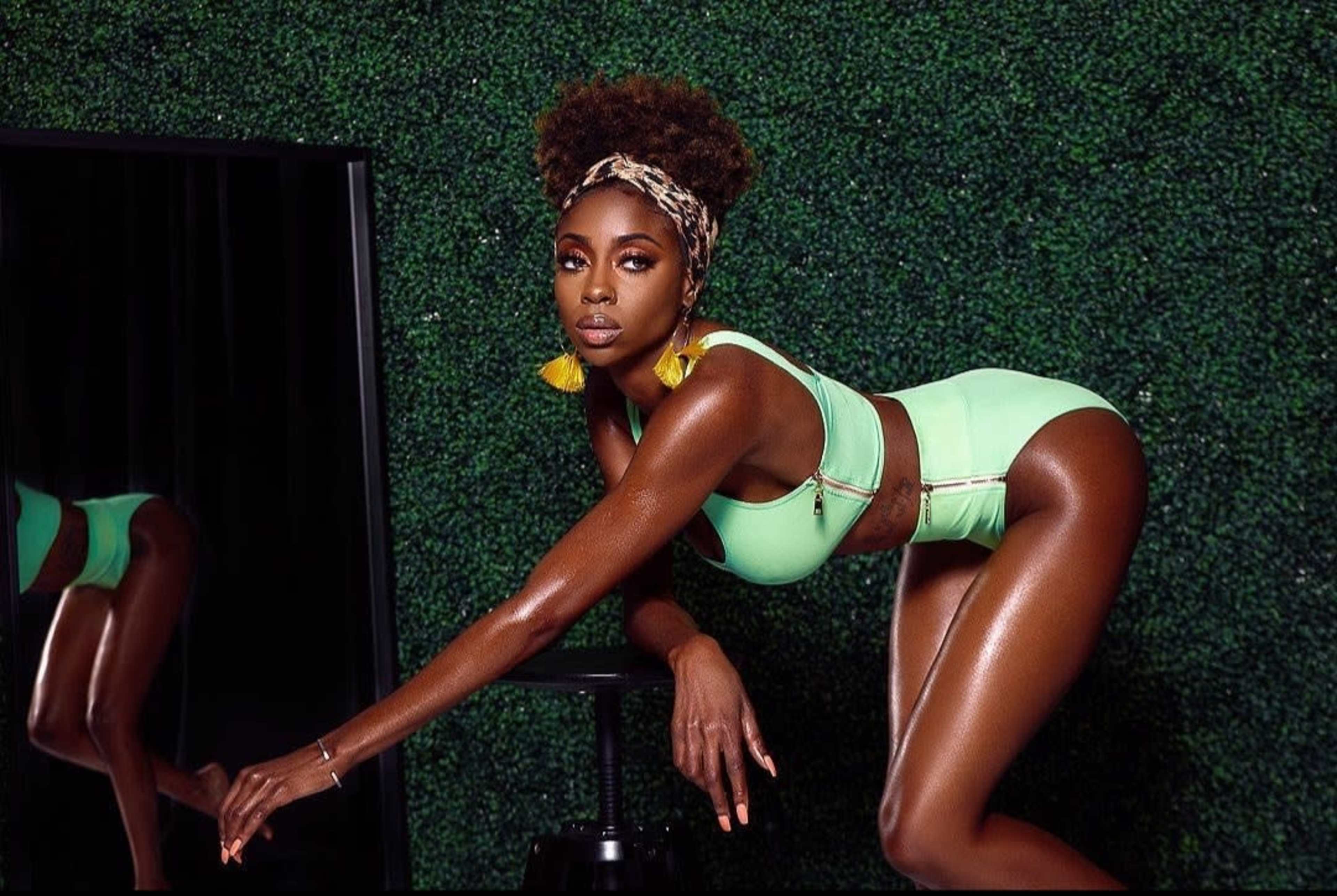 A model poses in a green two-piece outfit against a backdrop of artificial greenery while leaning on a black stool.