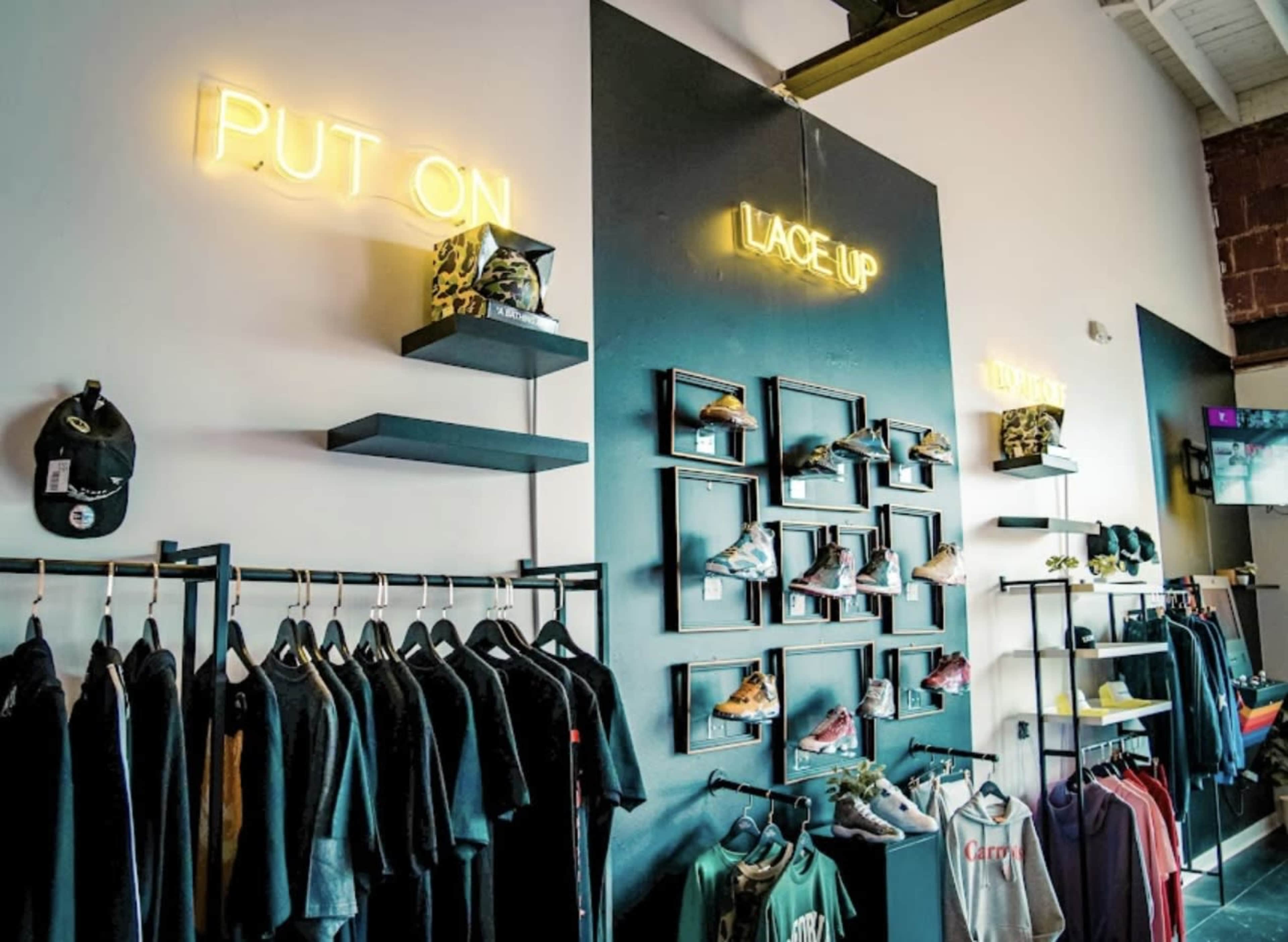 The image shows a retail display featuring clothing and footwear on racks and shelves, with neon signs reading "PUT ON" and "LACE UP" above the items.
