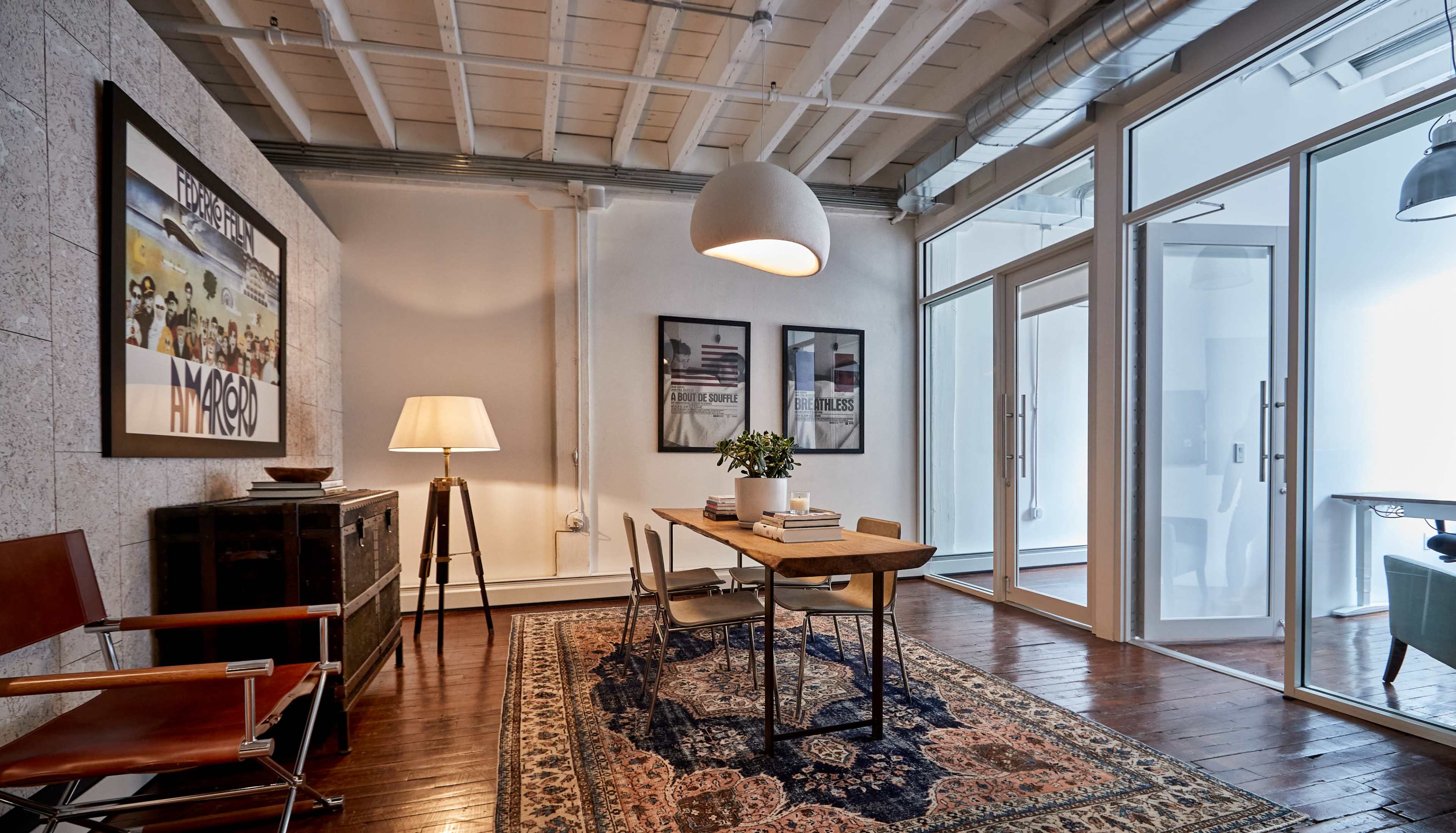 A modern workspace with a table and chairs, framed artwork on the wall, a floor lamp, and a patterned rug on hardwood flooring.