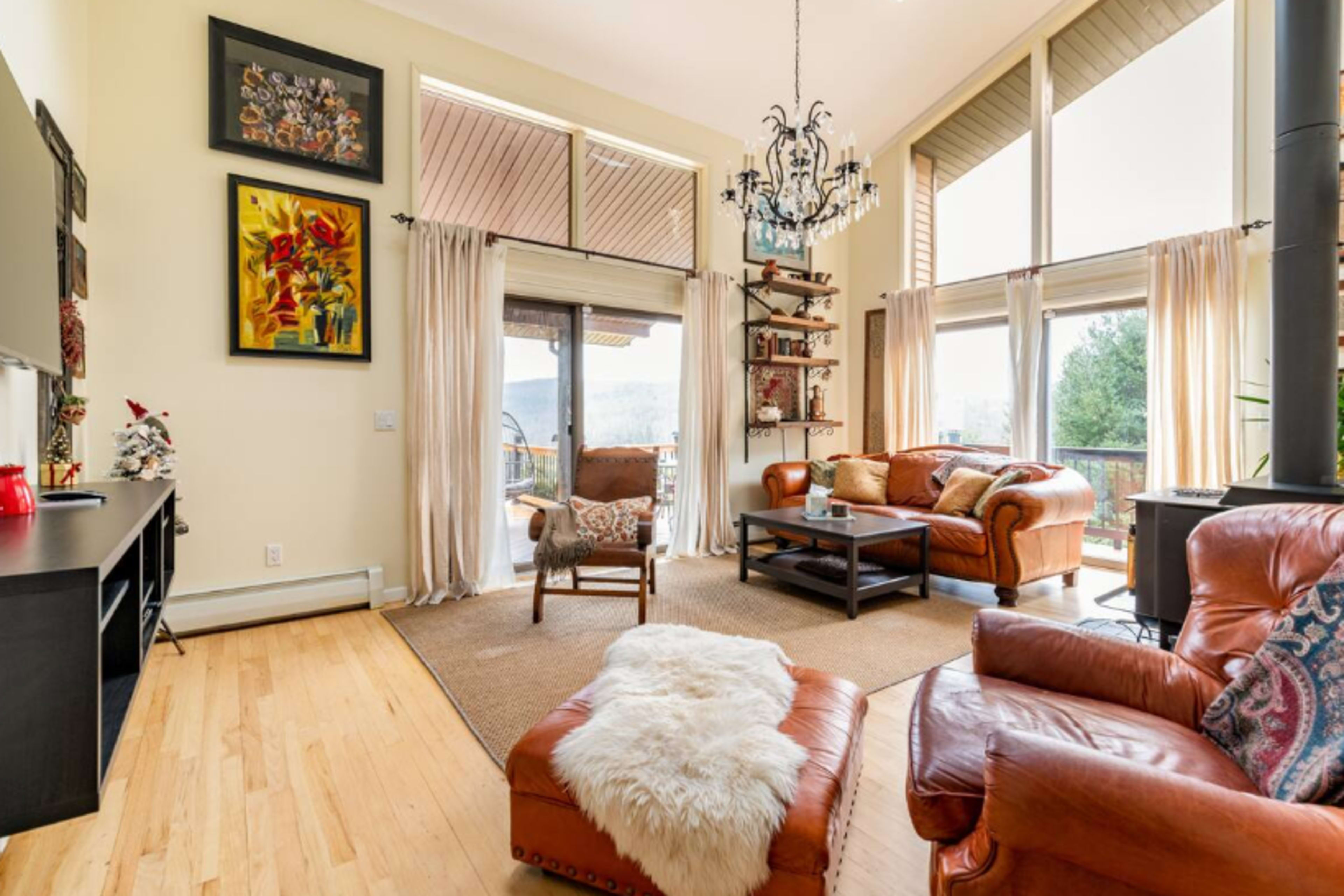 The living room features large windows, a chandelier, and a mix of leather seating arrangements with a wooden coffee table.