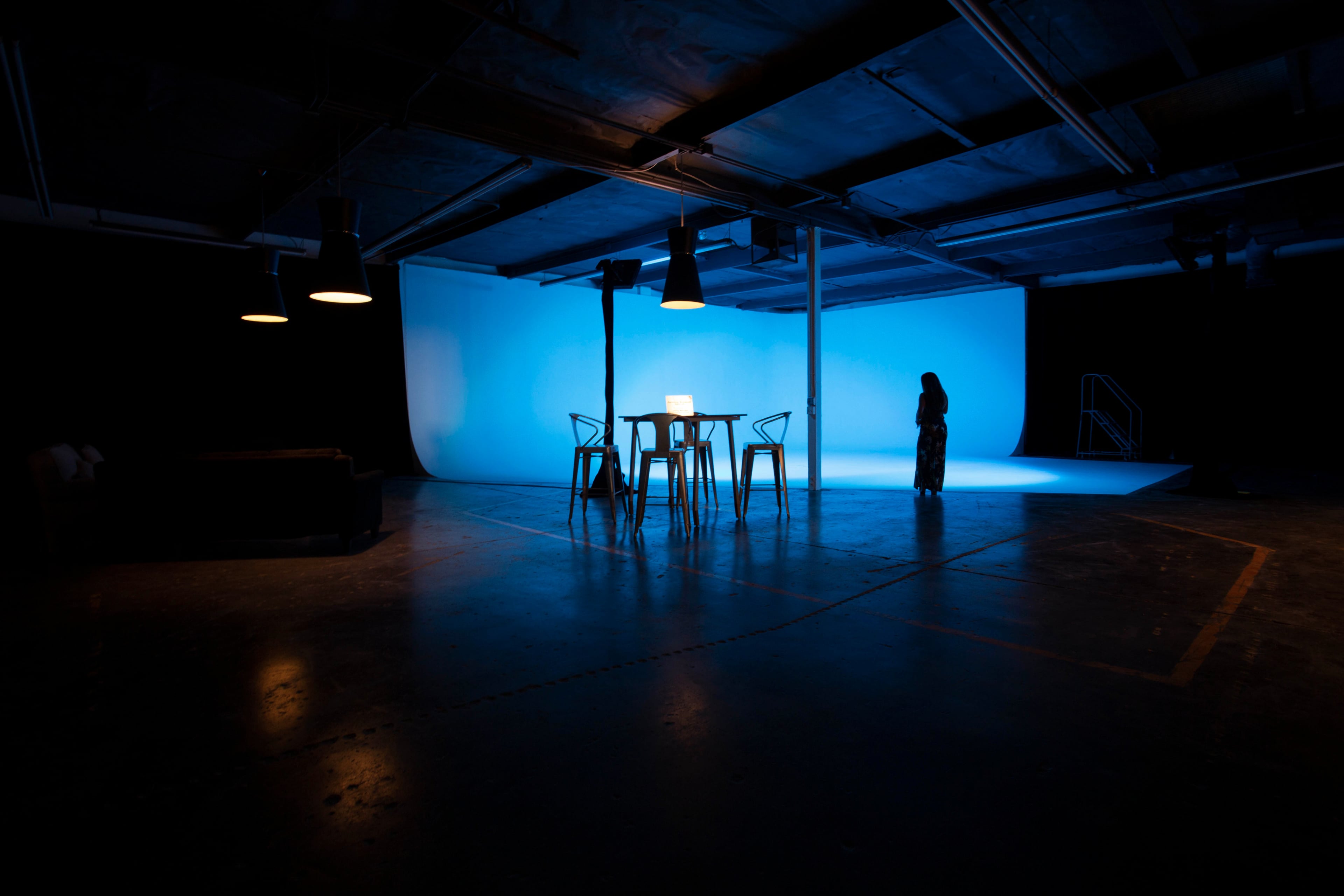 A dimly lit studio features a blue backdrop, a table with a laptop, and a solitary figure standing nearby.