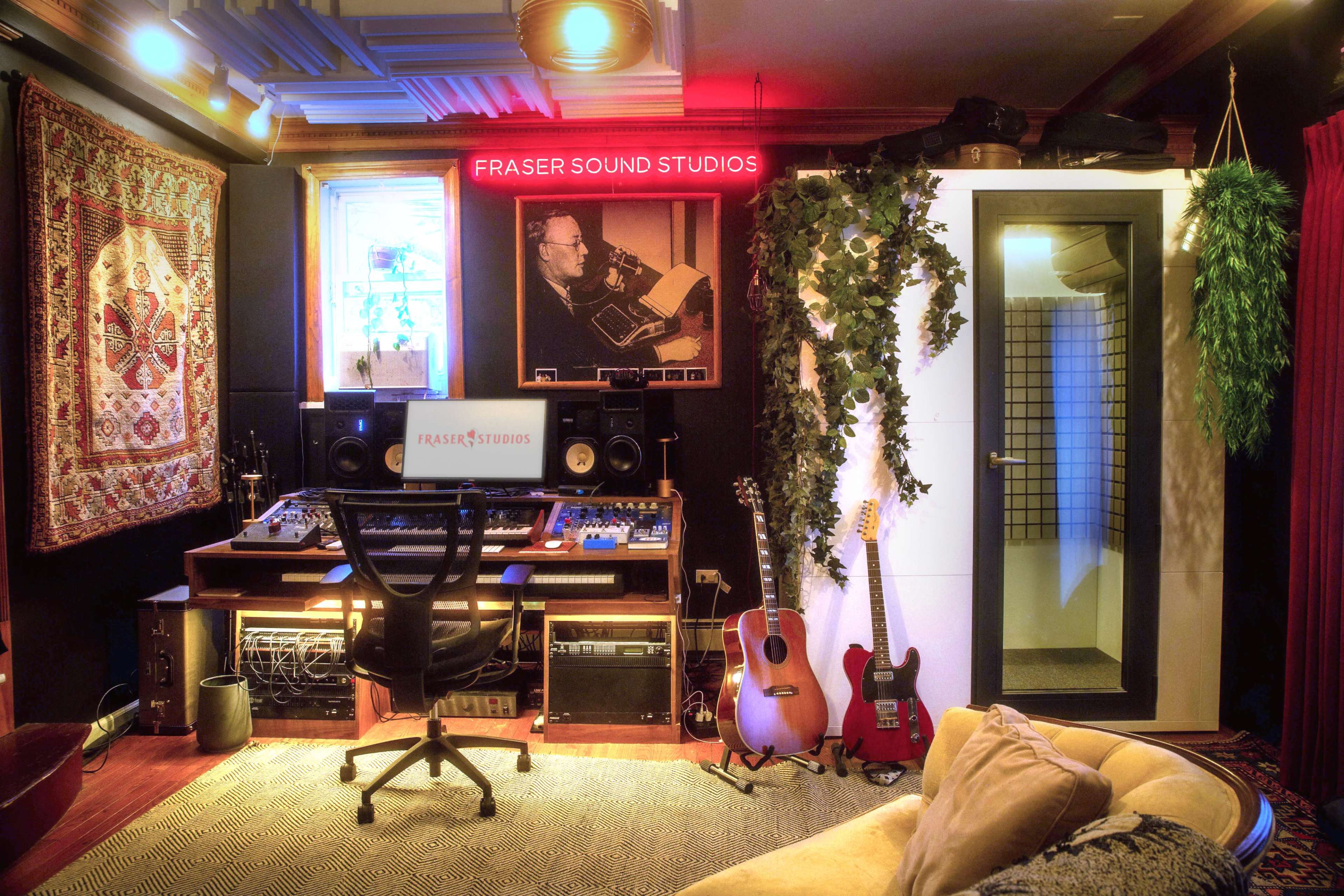 A music studio with a mixing desk, guitars, a vocal booth, and a large wall photo, all decorated with a tapestry and greenery.