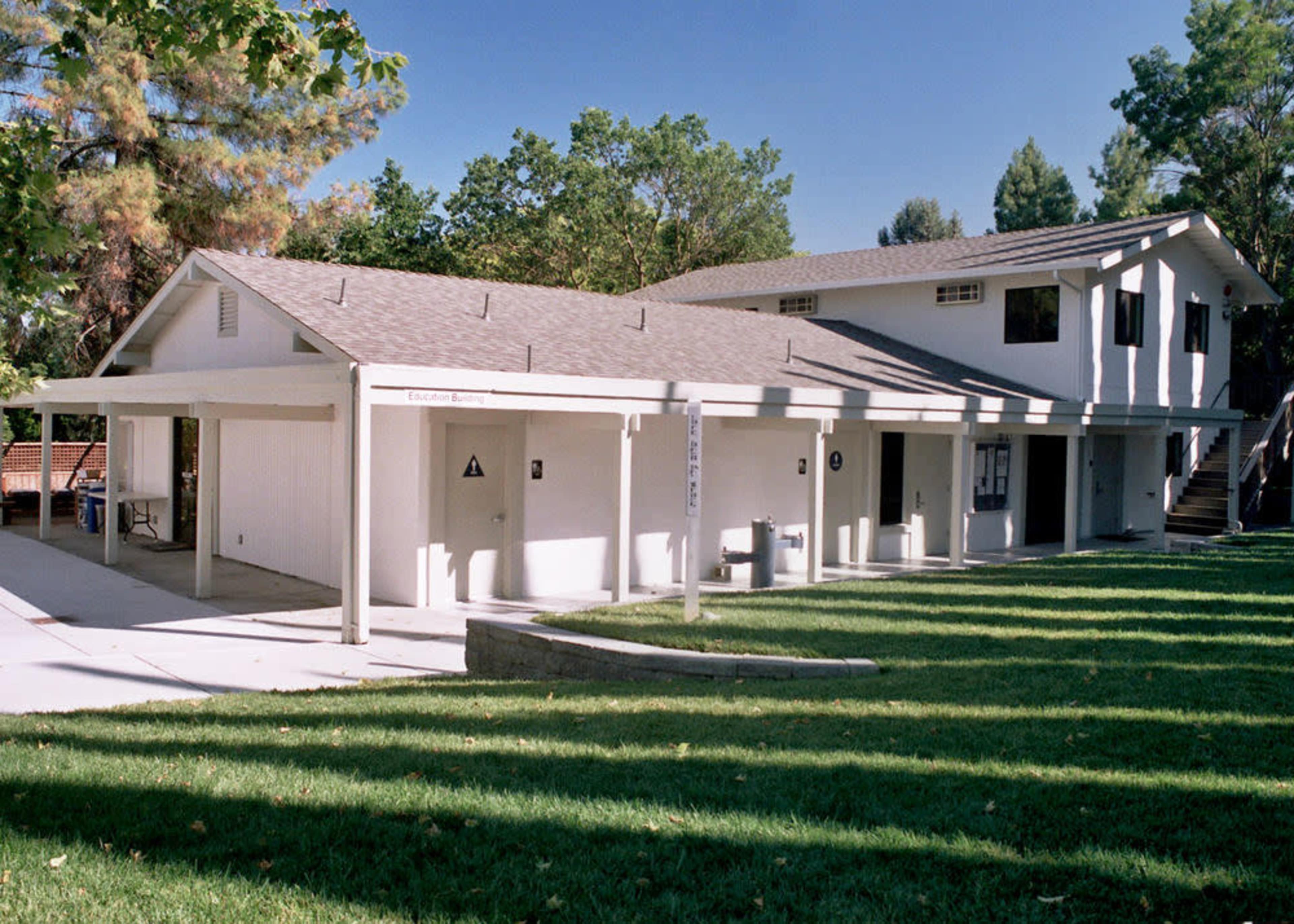 A two-story building with a sloped roof, situated next to a grassy area and surrounded by trees.