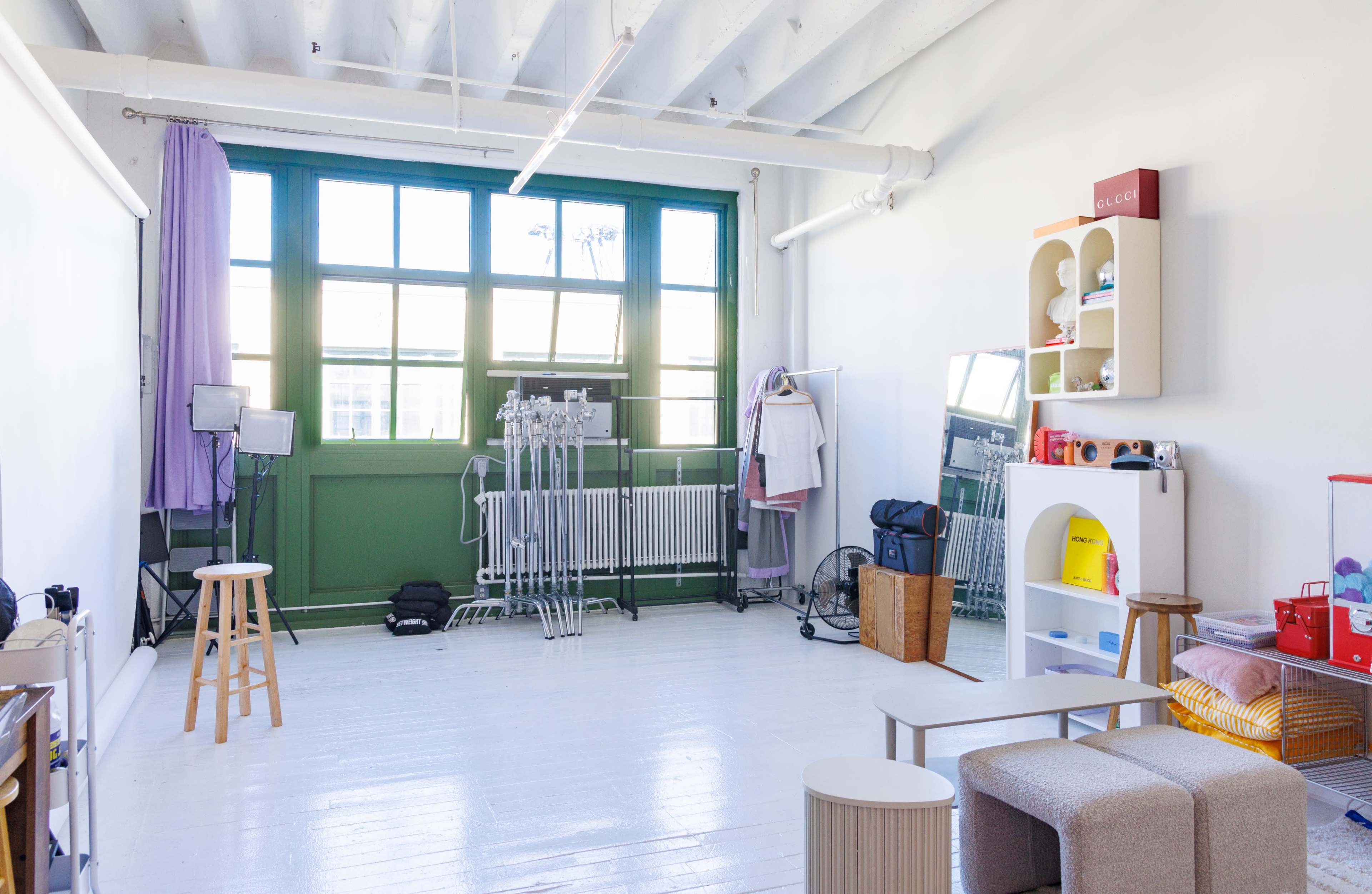 The image shows a spacious room with white walls and a hardwood floor, featuring various types of furniture, including stools, storage units, and lighting equipment, along with large windows letting in natural light.