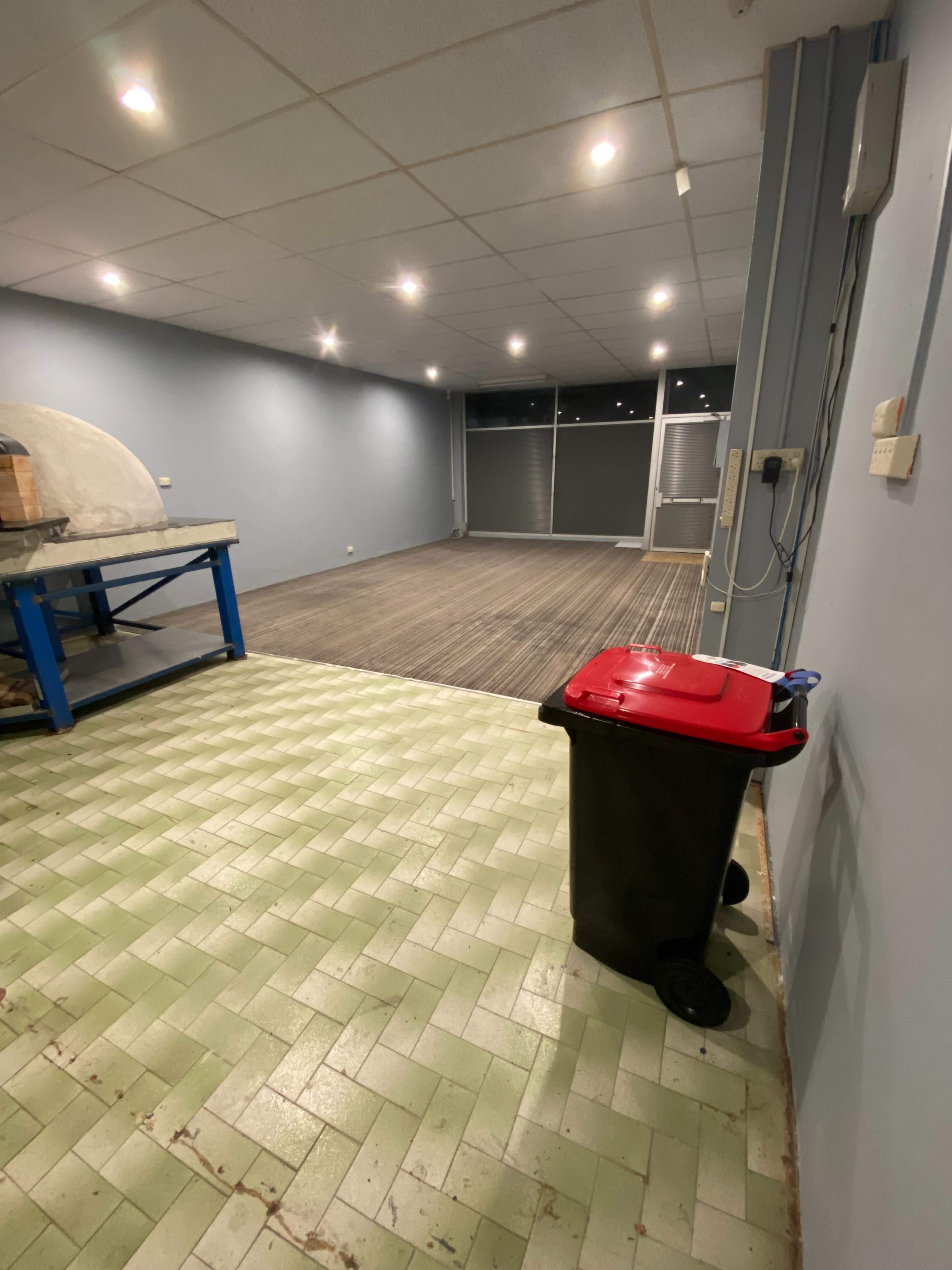 An empty room with light green and gray tiled flooring, a red-lidded trash bin, and blue tables along one wall.