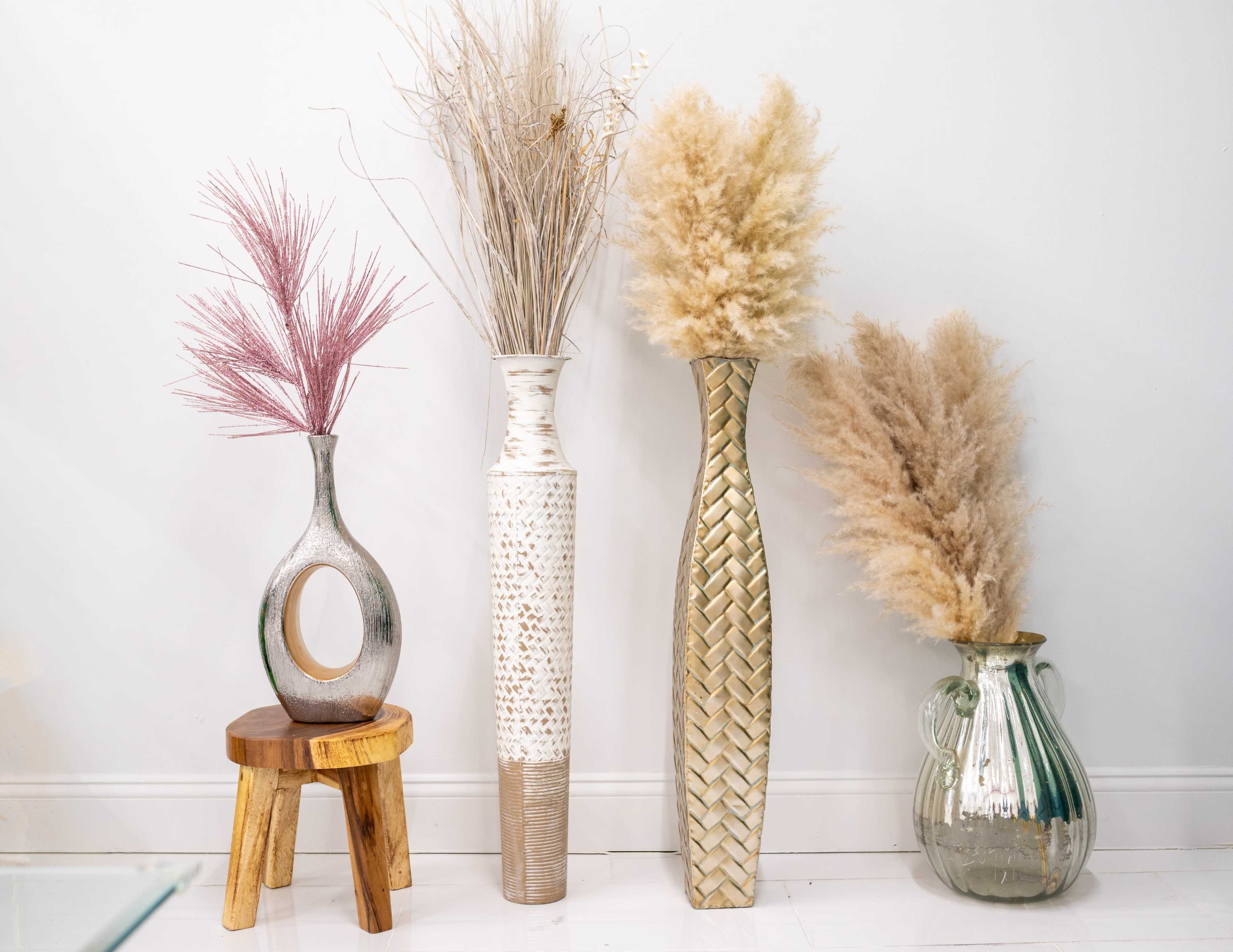 The image features four decorative vessels displaying various dried floral arrangements, including pampas grass, against a plain white wall.