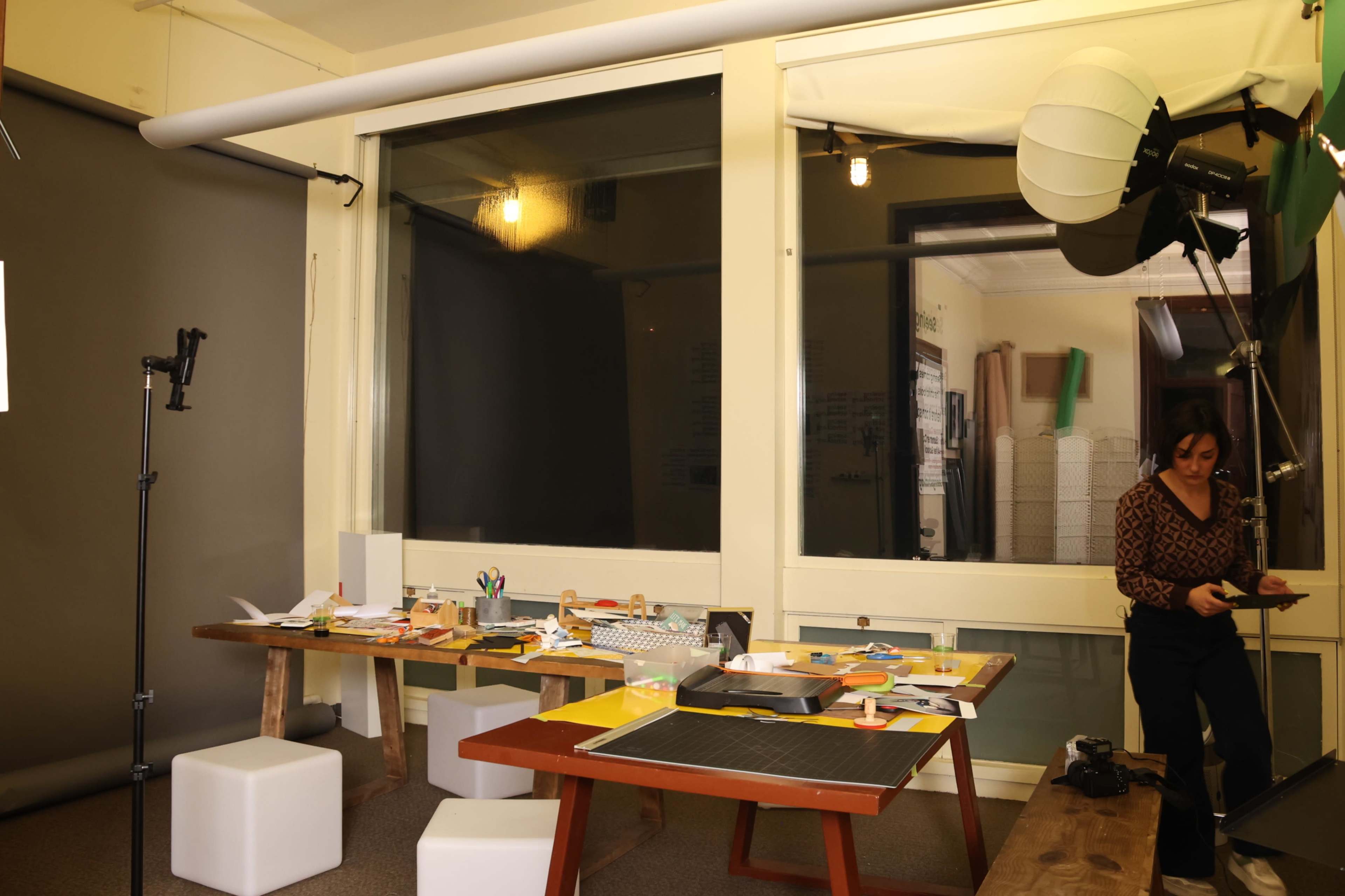 The image shows a workspace with tables covered in papers and craft materials, along with photography equipment set up in a well-lit room.