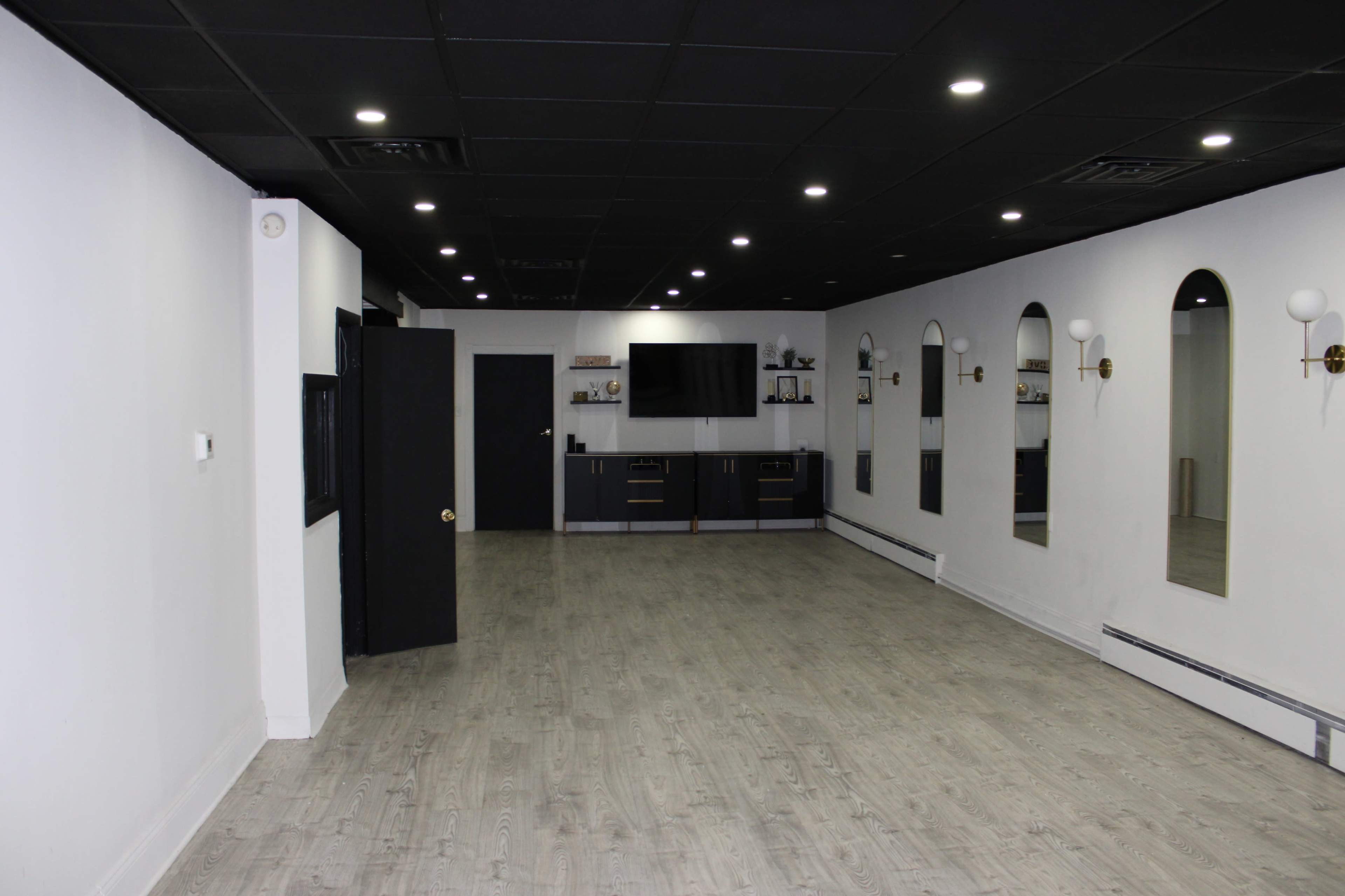 The room features a large, empty space with gray wood-like flooring, black ceiling, and white walls adorned with mirrors and wall sconces.