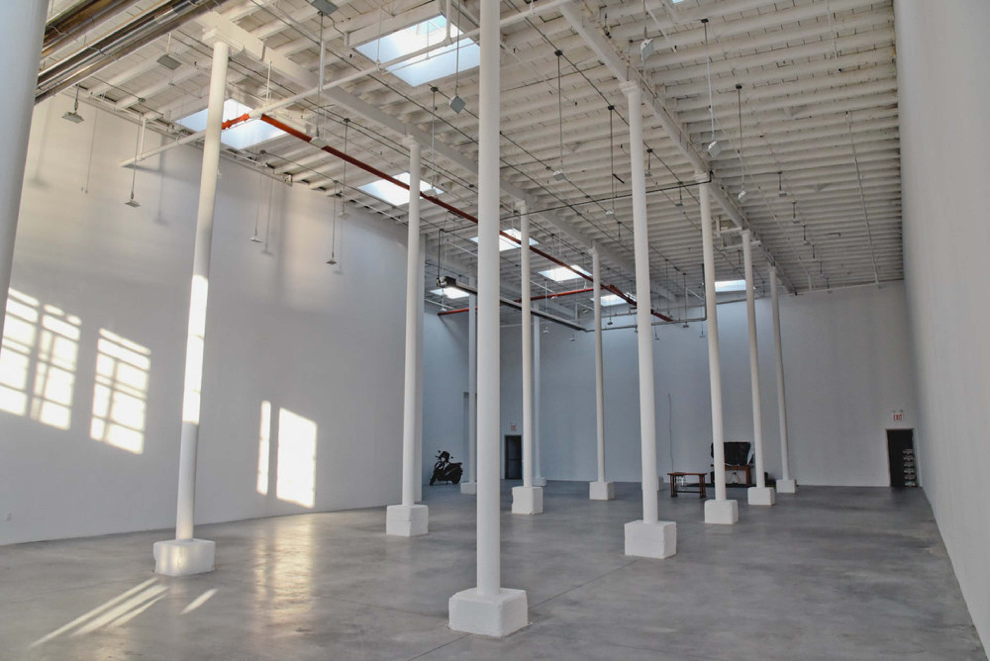 A spacious, empty interior with white walls and several tall columns, illuminated by natural light coming through skylights.