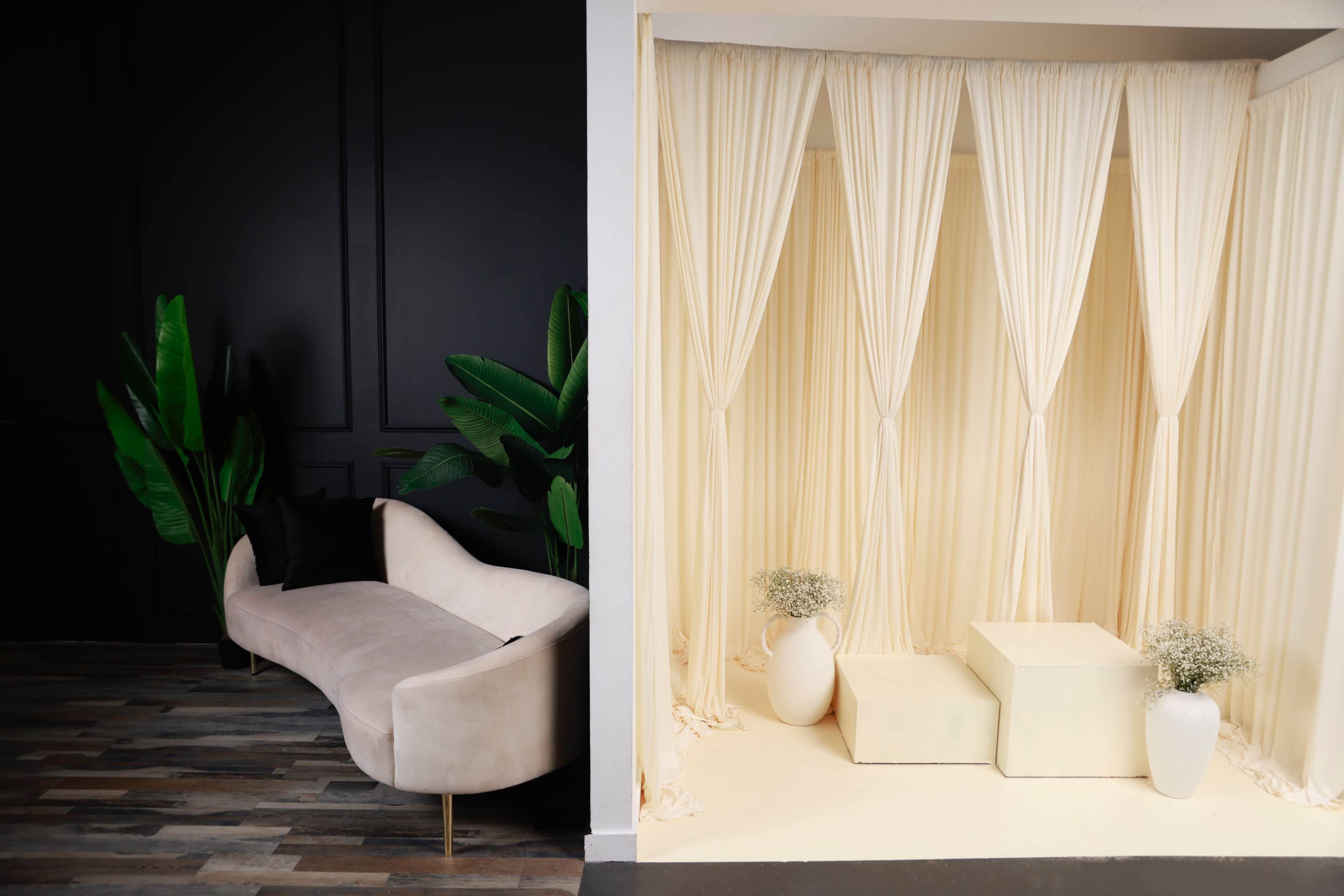 The image shows a split scene with a plush sofa on the left against a black wall and a small stage with cream curtains and two white pedestals on the right.