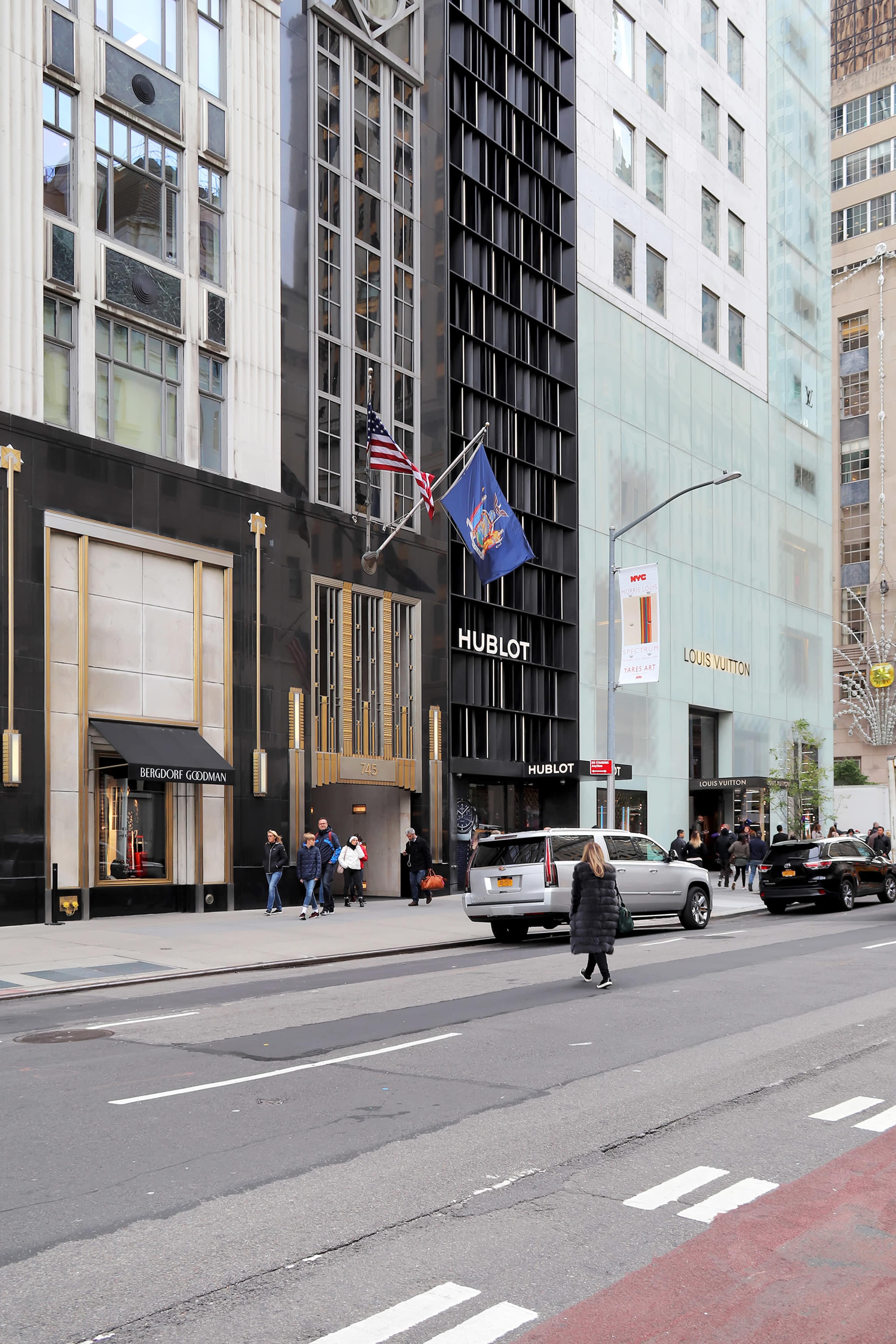 The image shows a street scene in an urban area with a Hublot store prominently displayed, flanked by a Louis Vuitton store and a flagpole featuring a state flag.