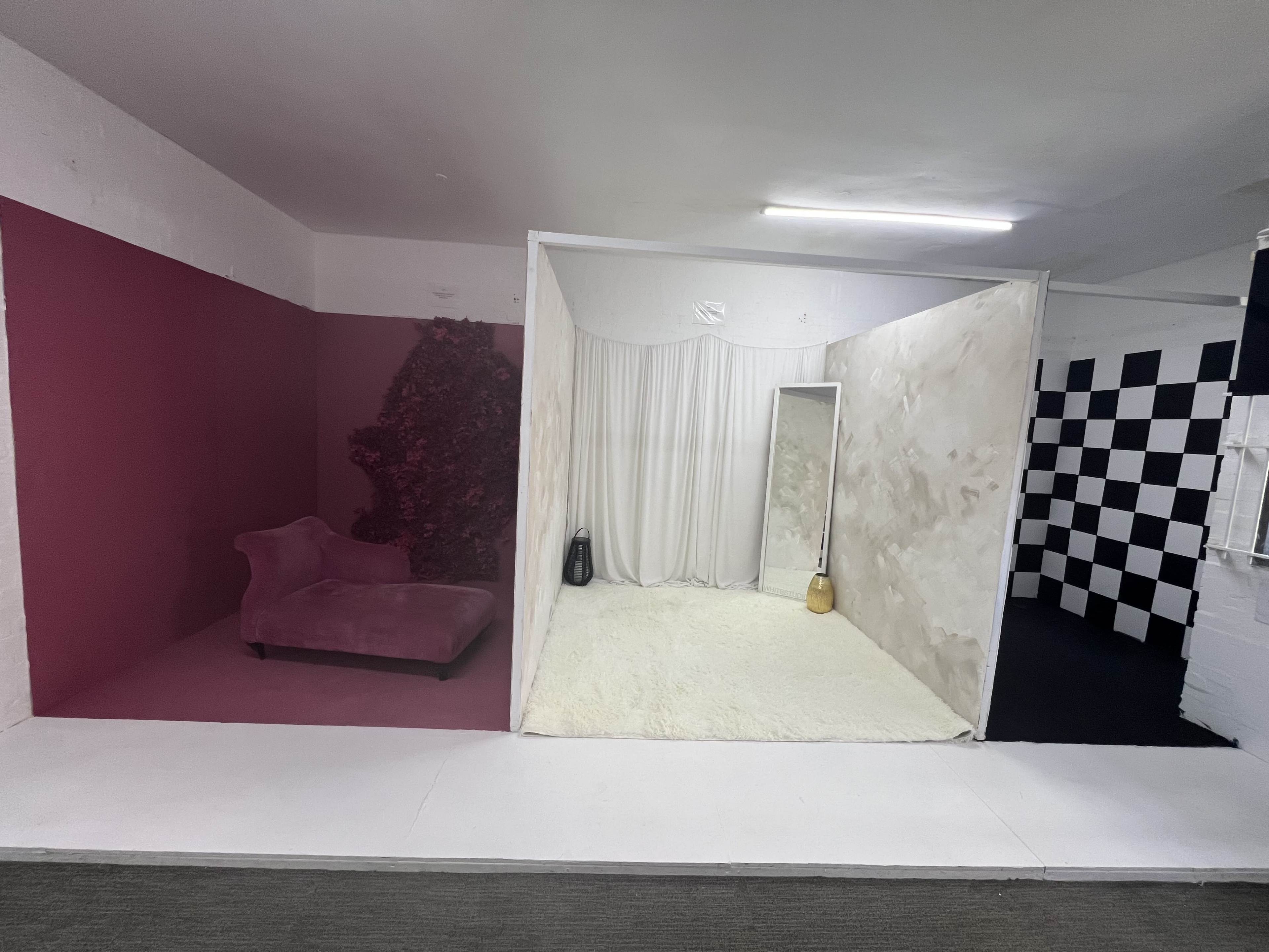 The image shows a divided space featuring a pink wall with a chaise lounge, a white area with a mirror and draped fabric, and a black and white checkered wall.