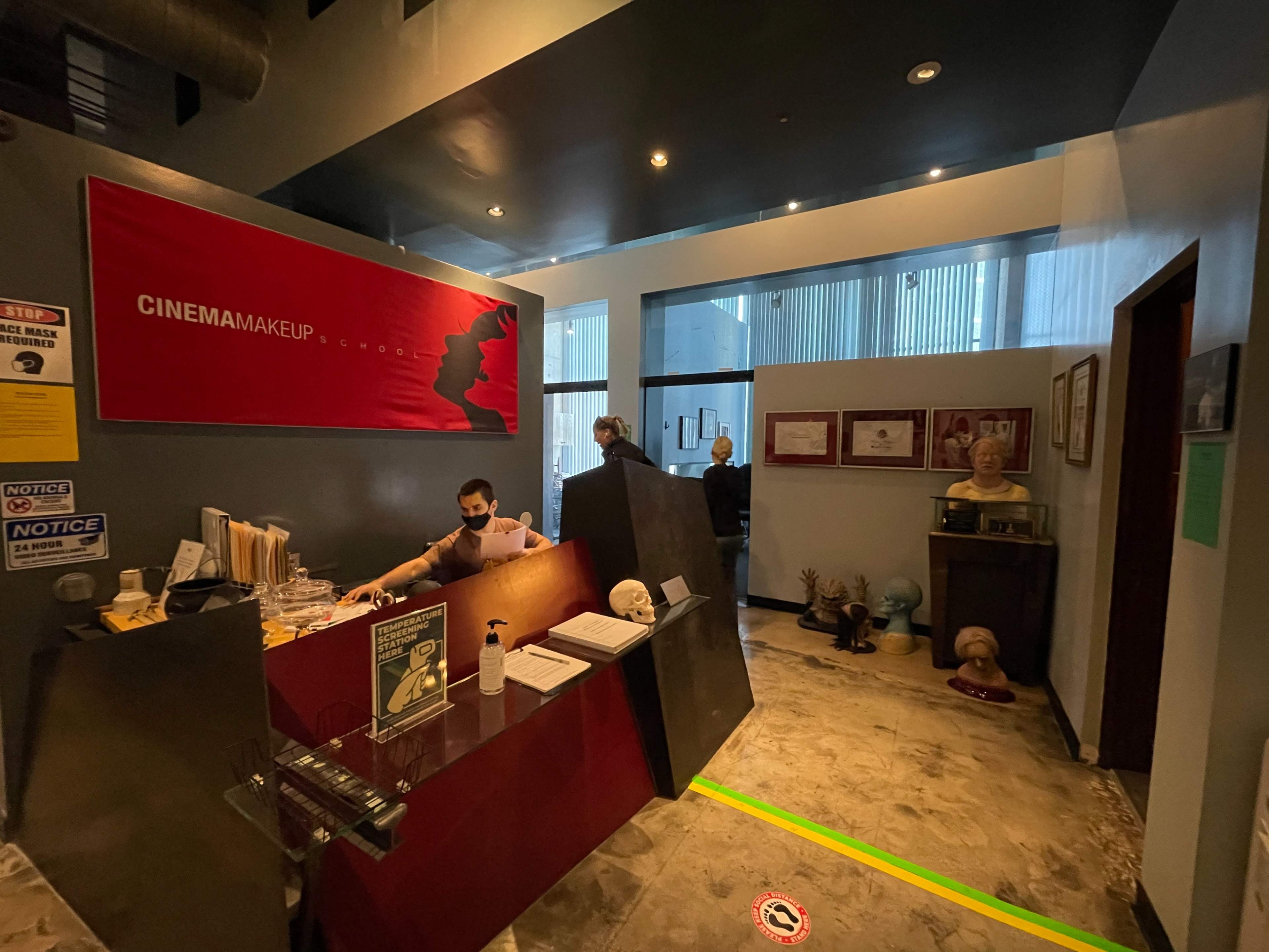 The image shows a makeup studio reception area with a red wall displaying "CINEMA MAKEUP," a receptionist at the desk, and a hallway leading to additional space.