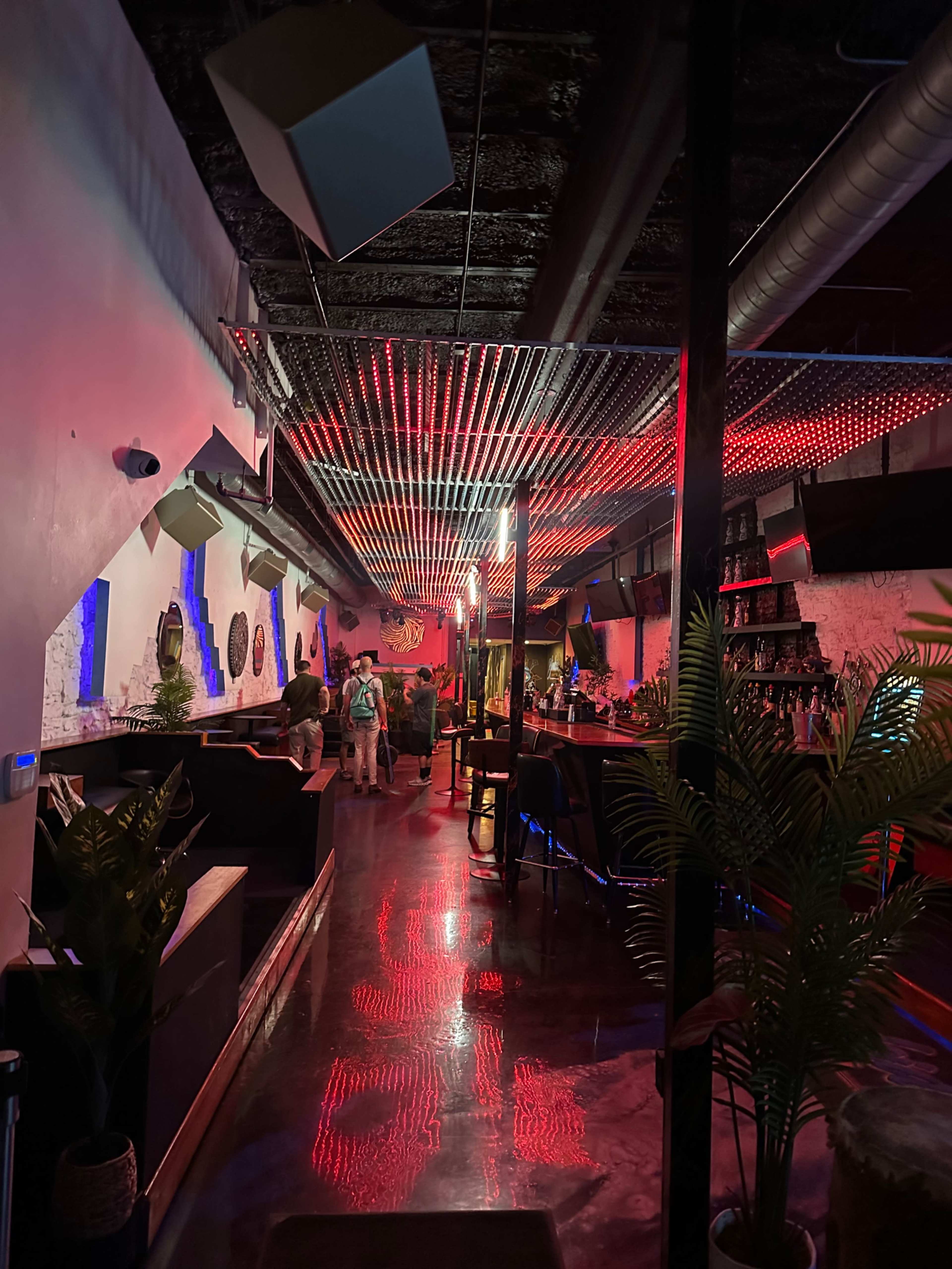 The image depicts an illuminated lounge with a long bar, featuring red and blue lighting reflecting off the polished floor.