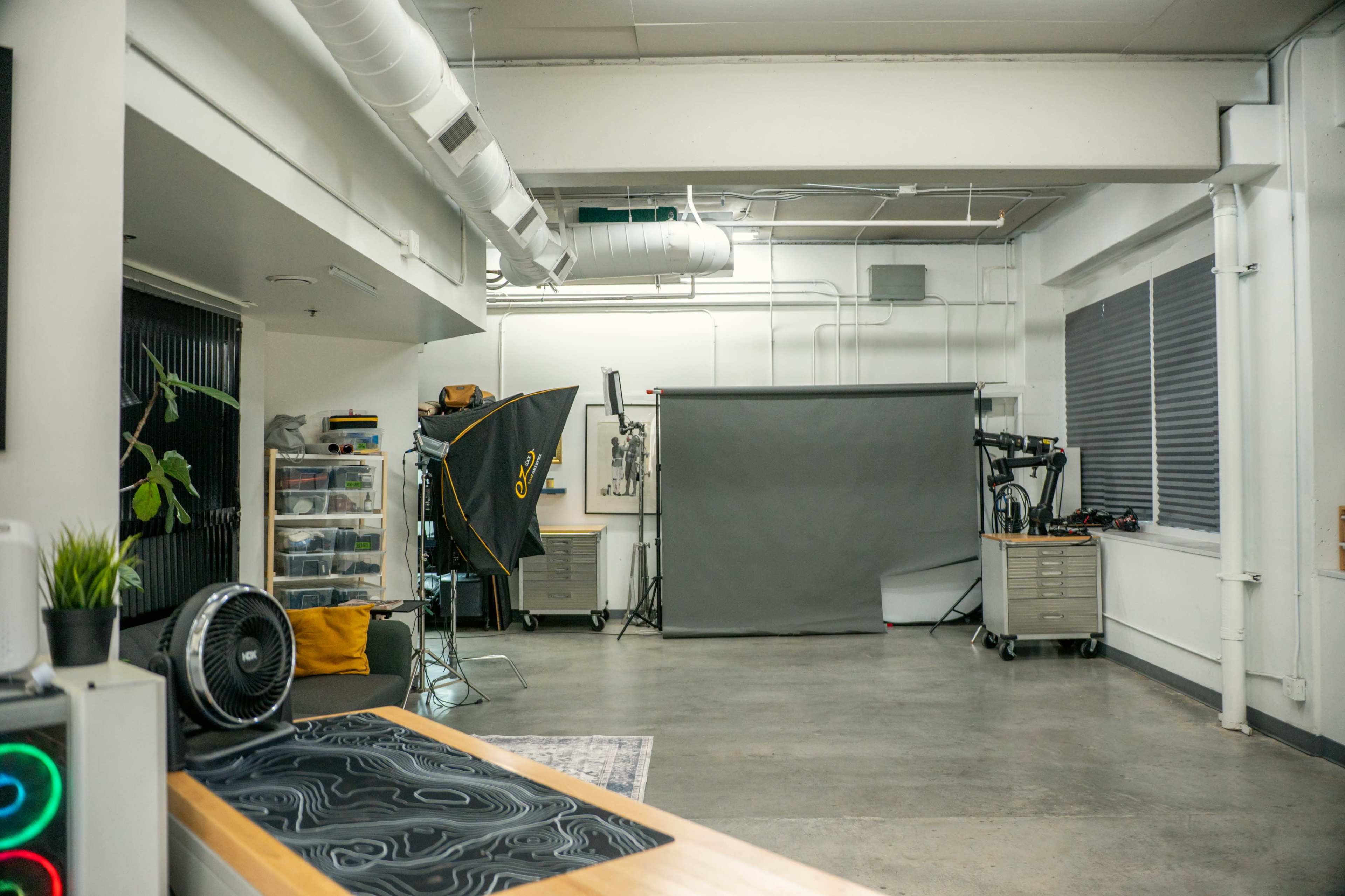 The image shows a spacious studio with a grey backdrop, photography equipment, storage units, and a large table.