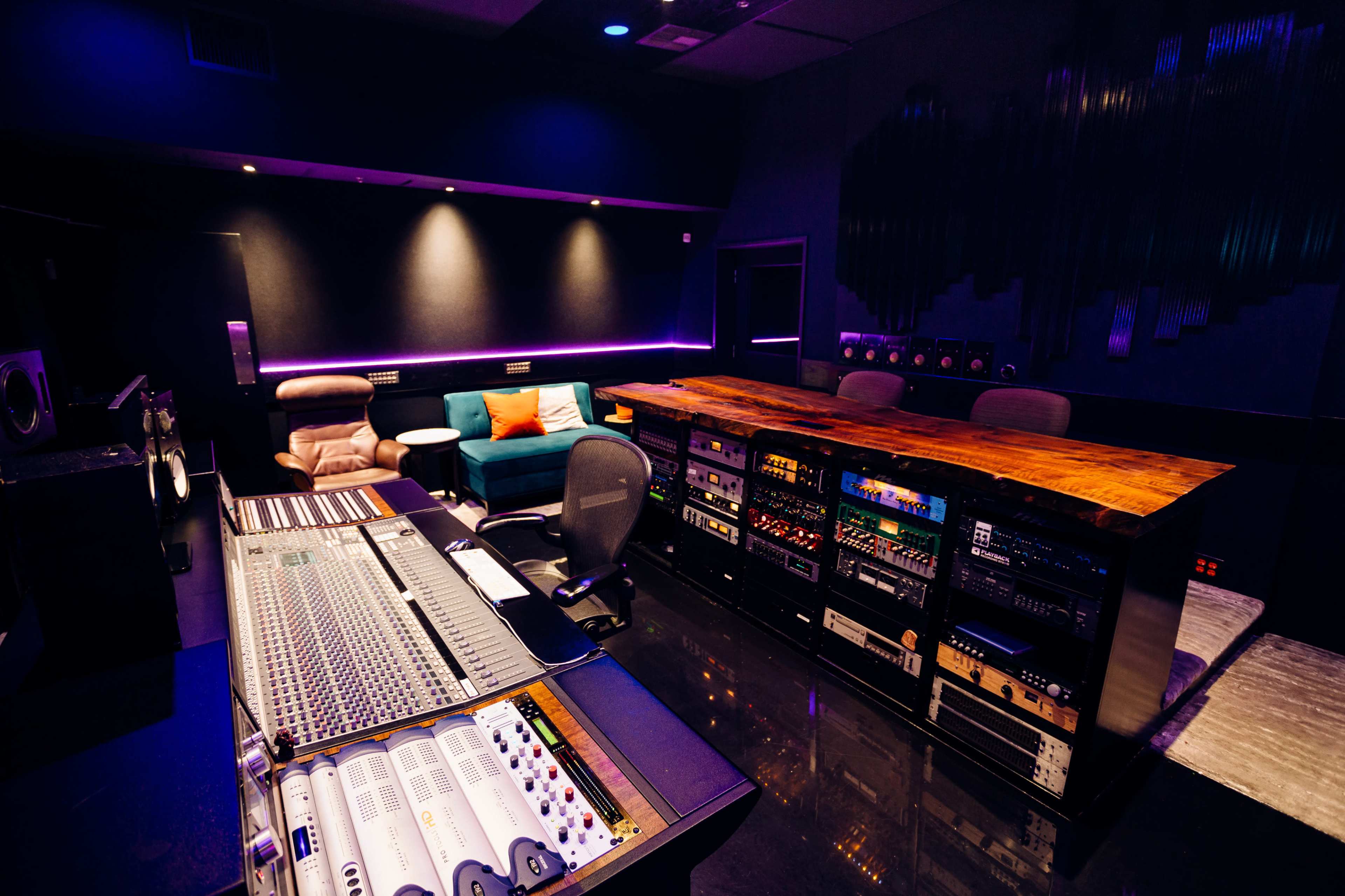 A modern recording studio featuring a large mixing console, a wooden work surface, and a lounge area with a teal sofa and orange pillows.