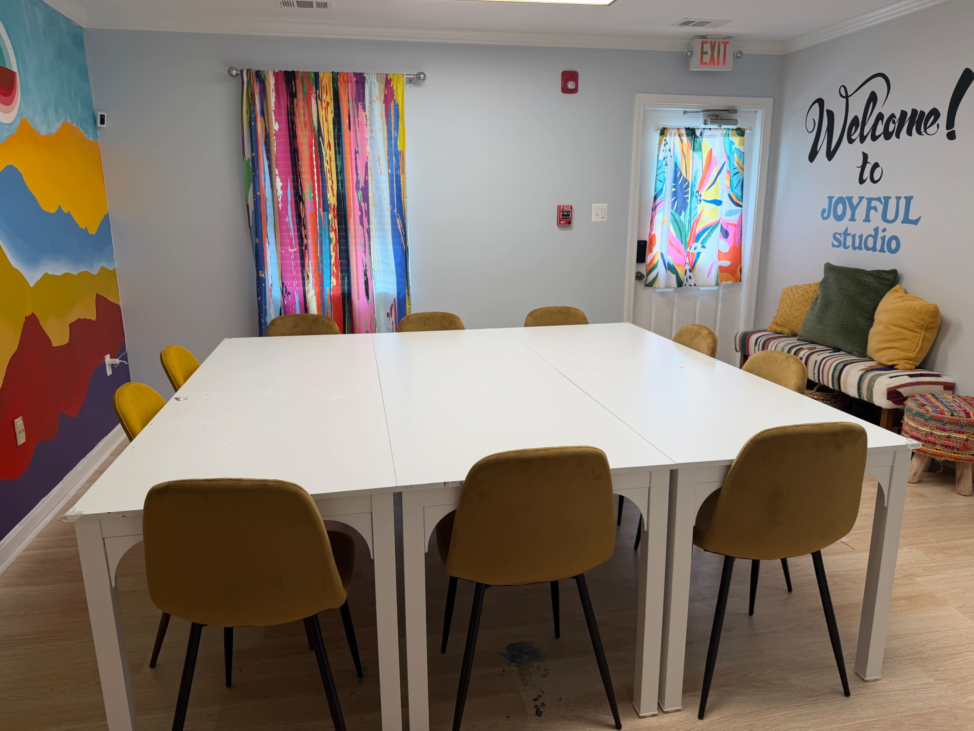 The image shows a brightly lit studio with a large white table surrounded by yellow chairs, colorful curtains, and a painted mural on one wall.