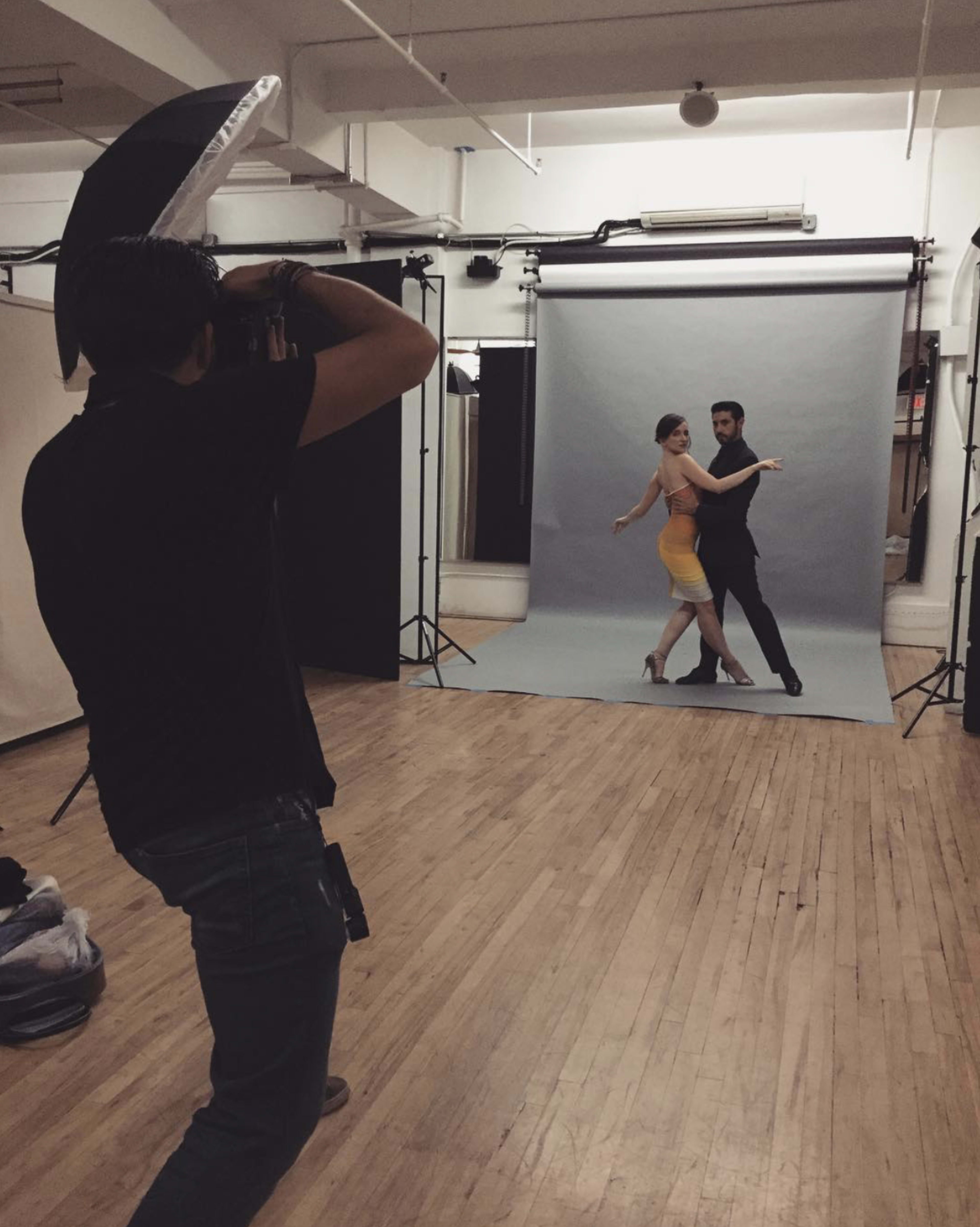 A photographer captures a couple dancing in front of a gray backdrop in a studio with wooden flooring.