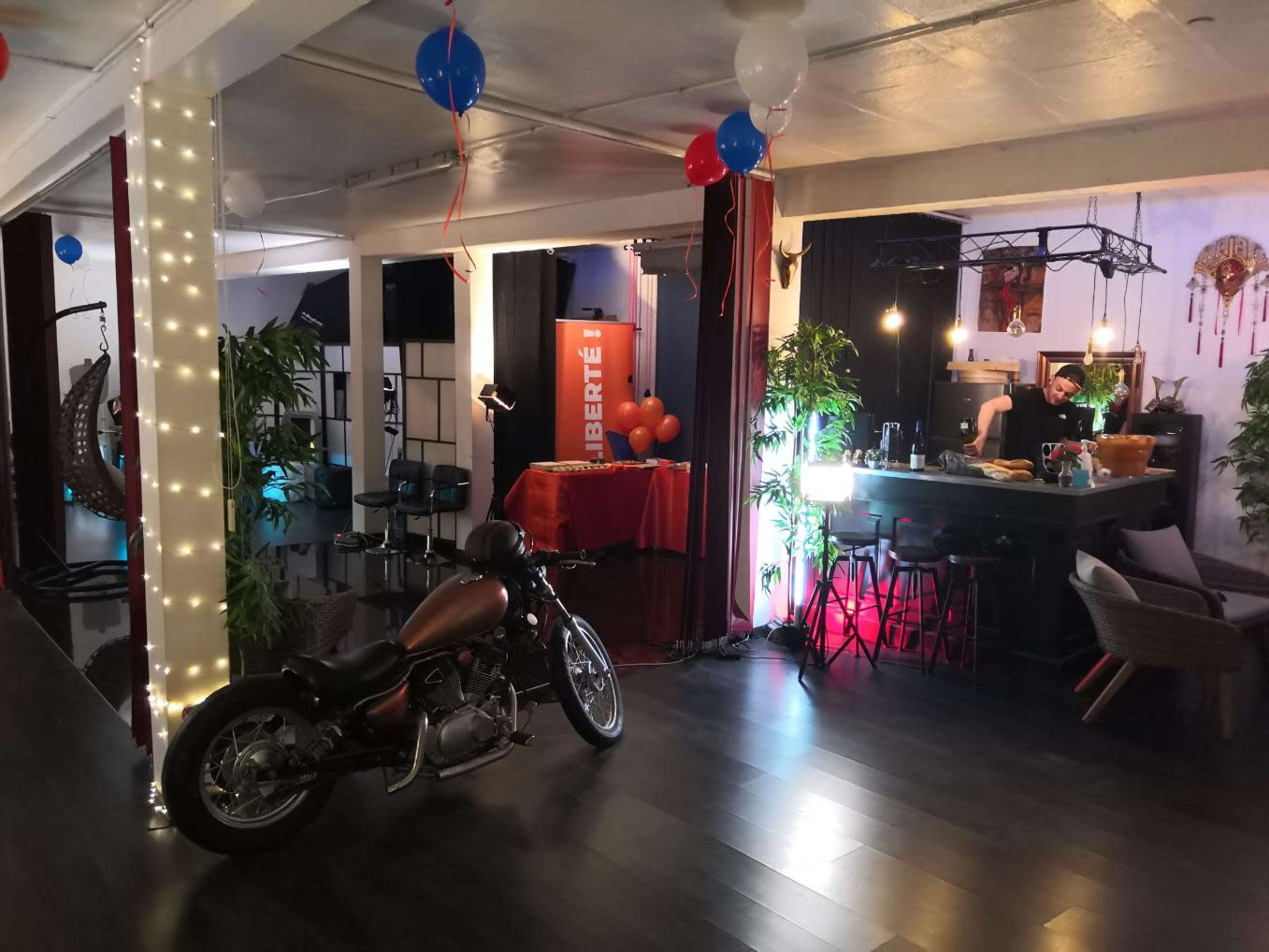 The image shows a decorated indoor space featuring a motorcycle, a bar setup with lights, and festive balloons.