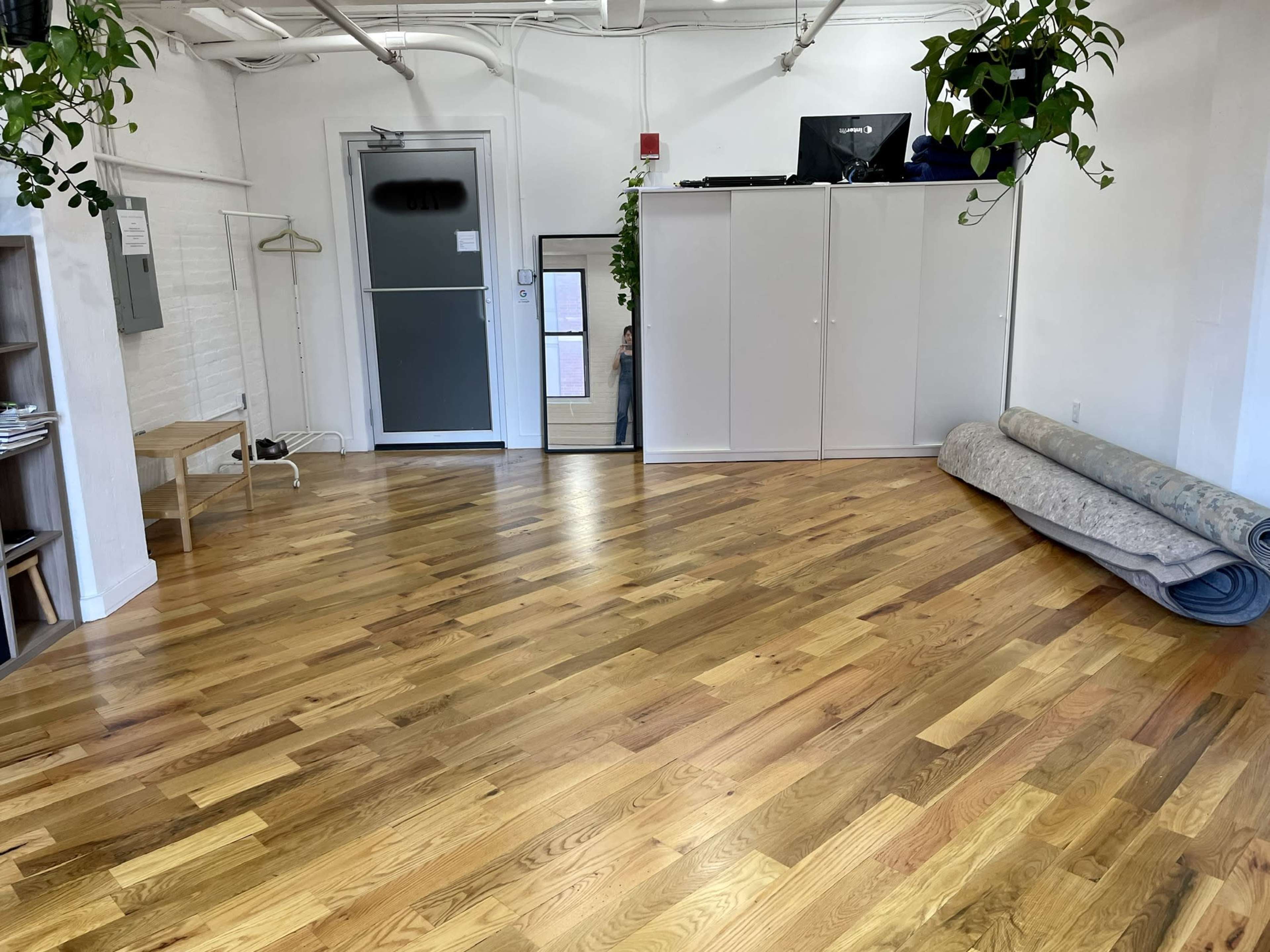 A spacious room with hardwood floors, a large mirror, a white cabinet, a stack of rolled-up carpet, and plants hanging from the ceiling.
