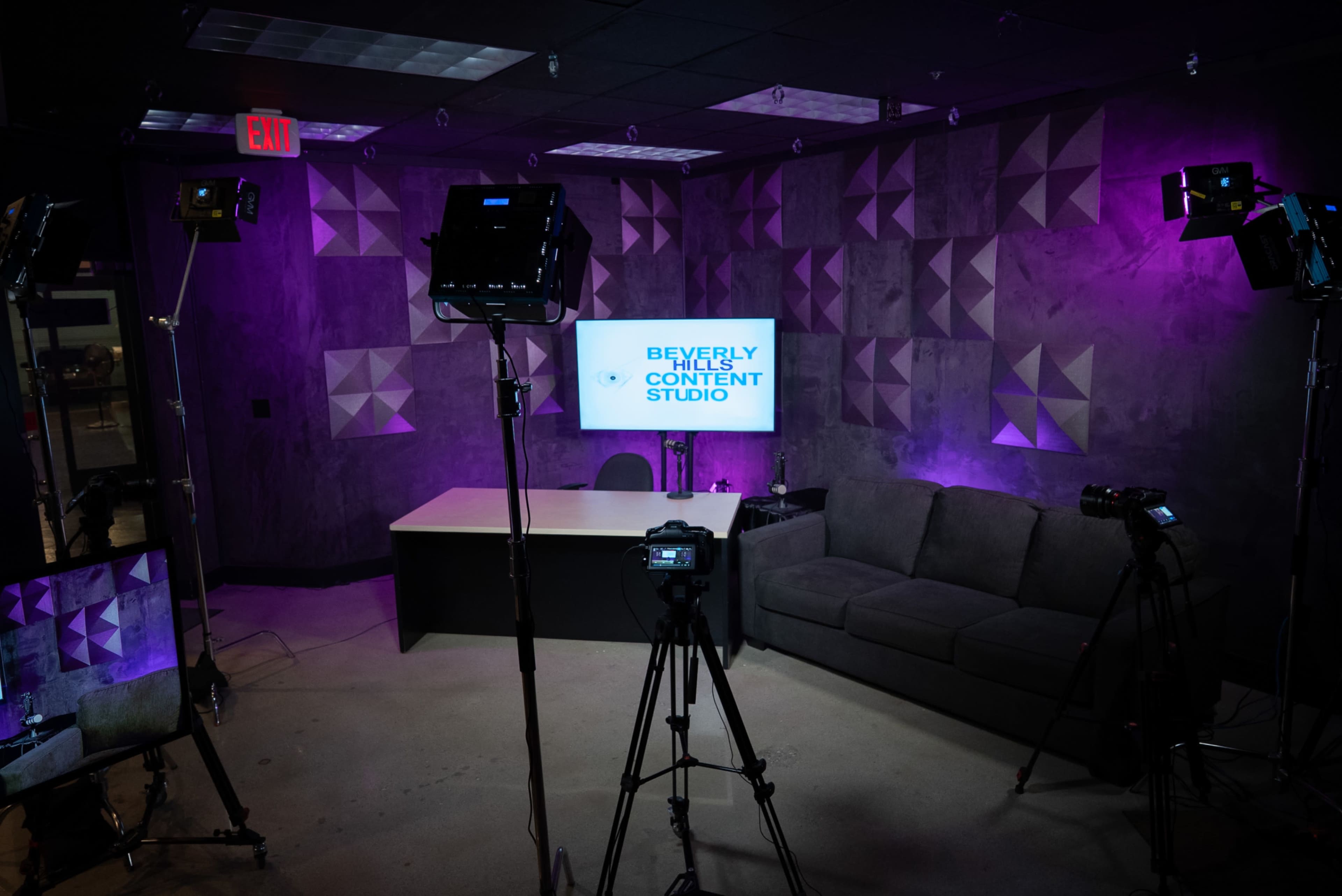 The image shows a modern content studio with a grey couch, a desk, and multiple camera setups facing a screen displaying "BEVERLY HILLS CONTENT STUDIO."