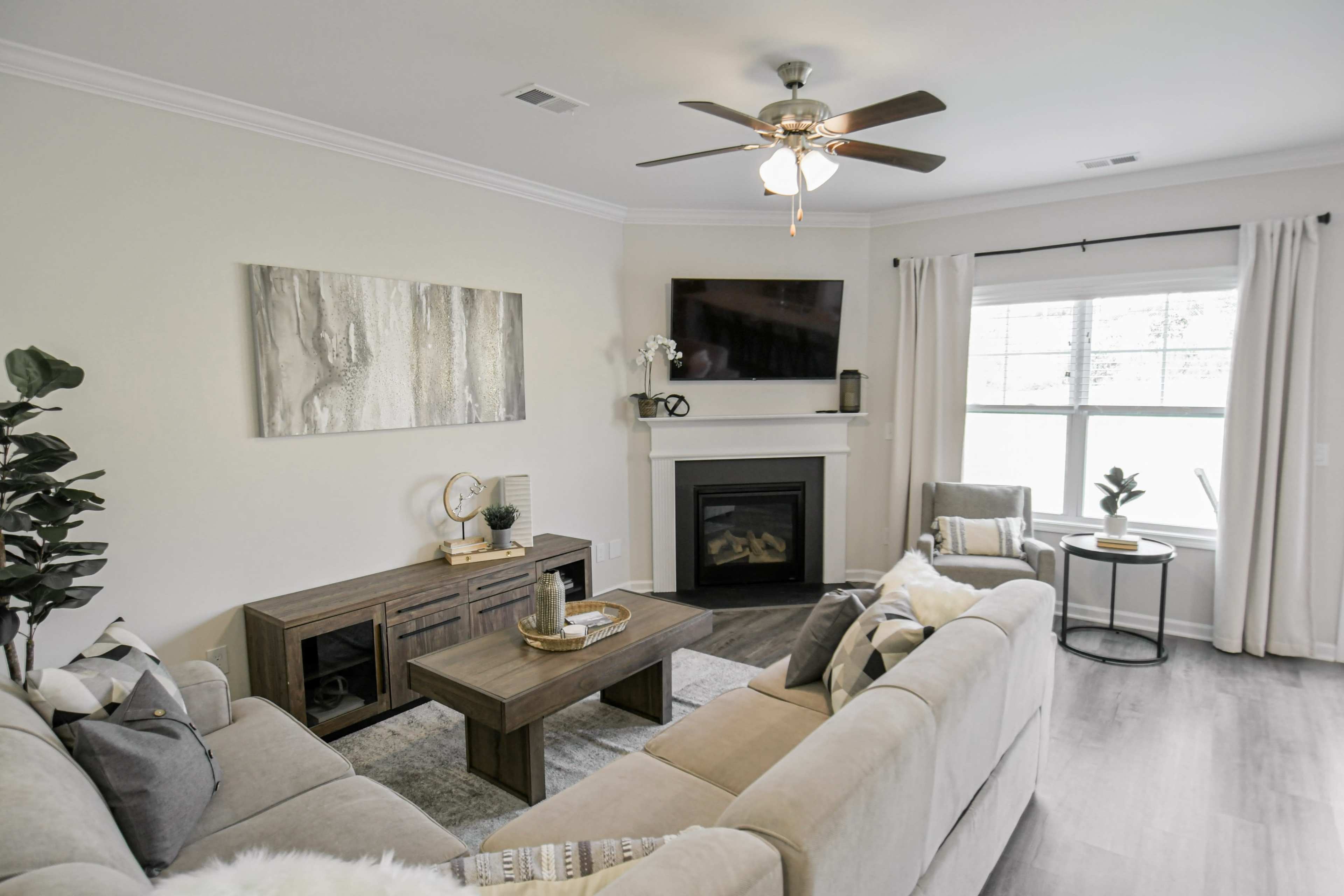 A modern living room features a beige sectional sofa, a wooden coffee table, and a fireplace, with large windows covered by light curtains.