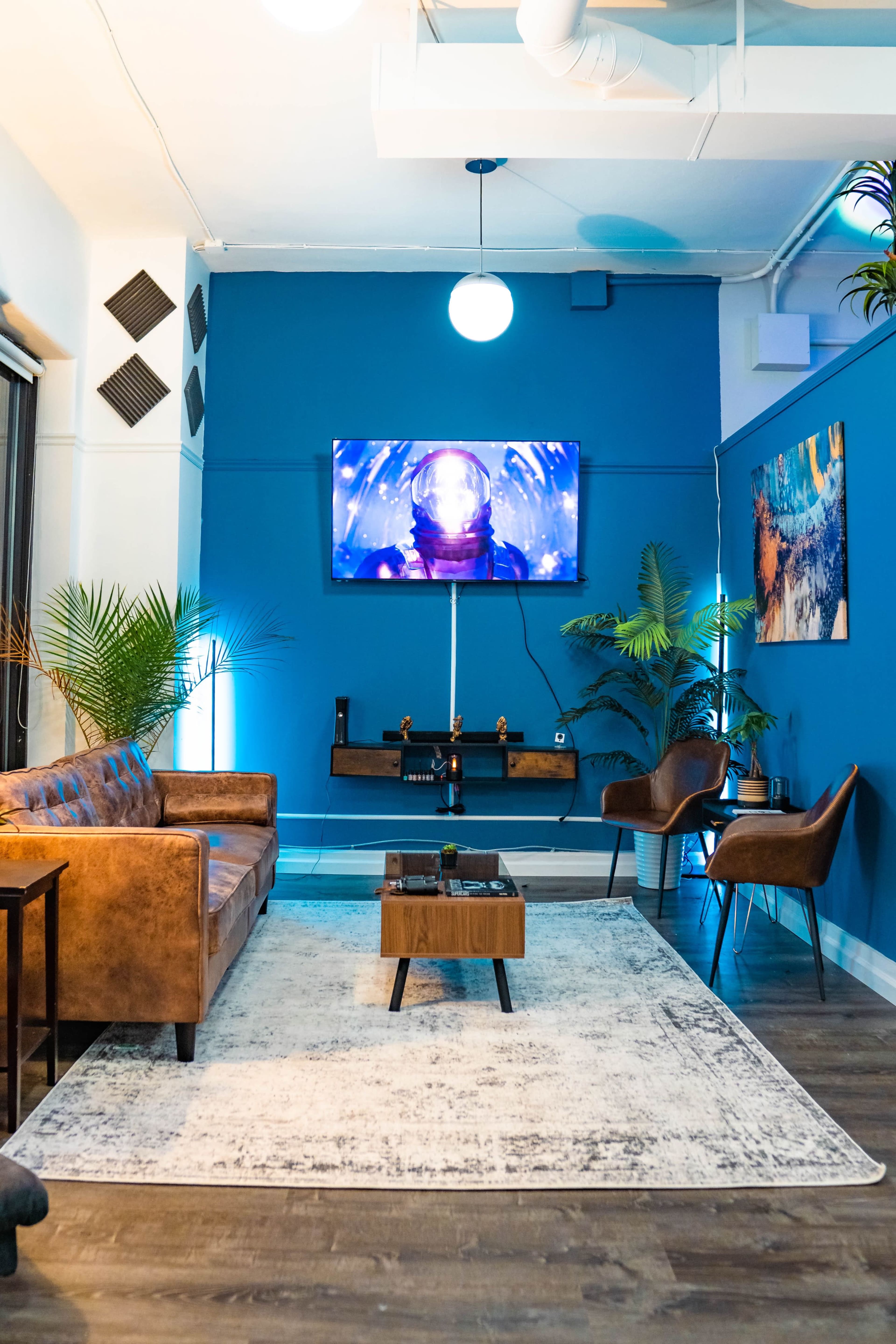 A modern living room with a brown leather sofa, two chairs, a wooden coffee table, and a blue wall featuring a television displaying a space-themed scene.