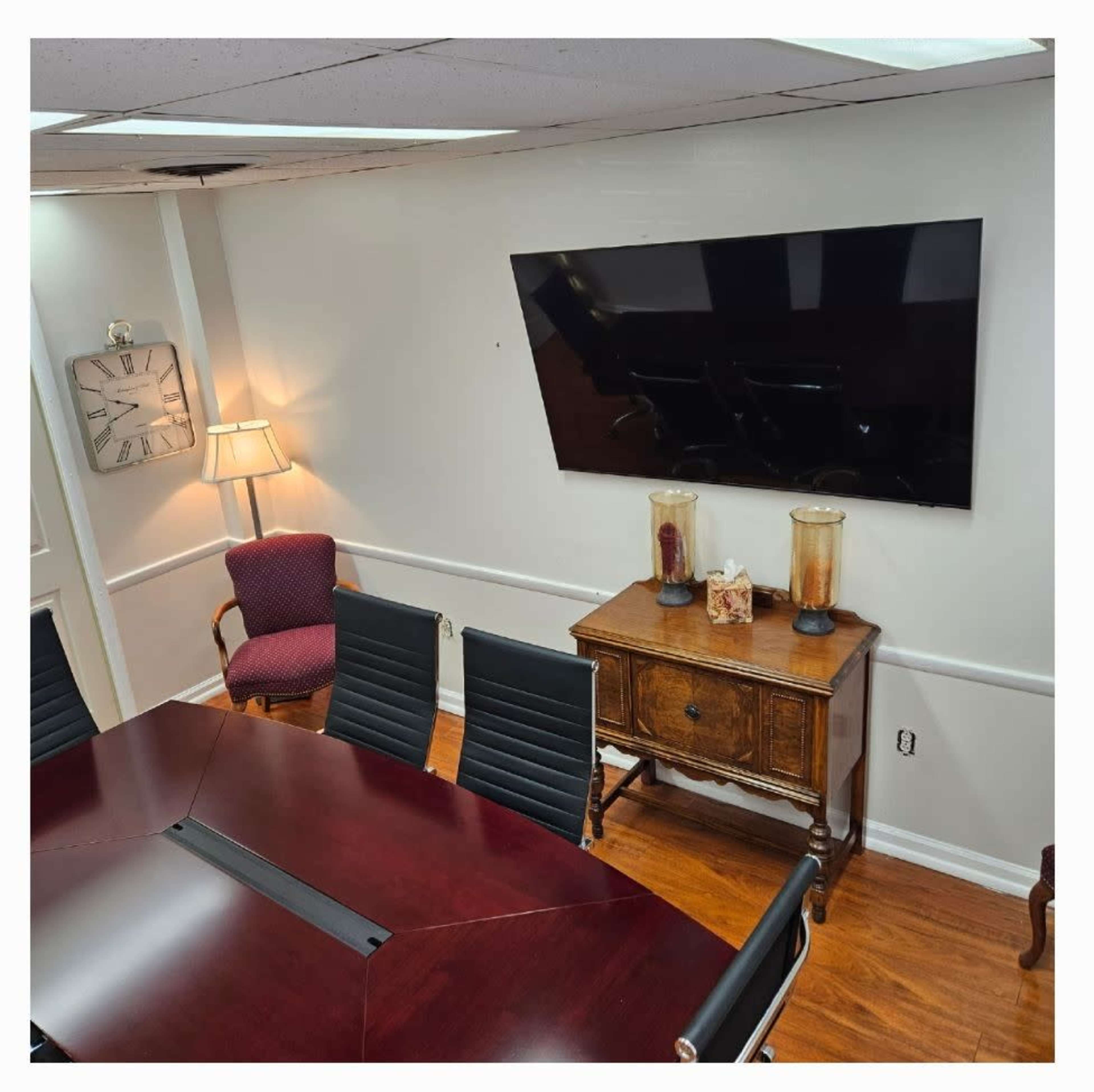 The image shows a conference room with a large table, black chairs, a wall-mounted TV, a standing lamp, and a wooden side table decorated with vases.