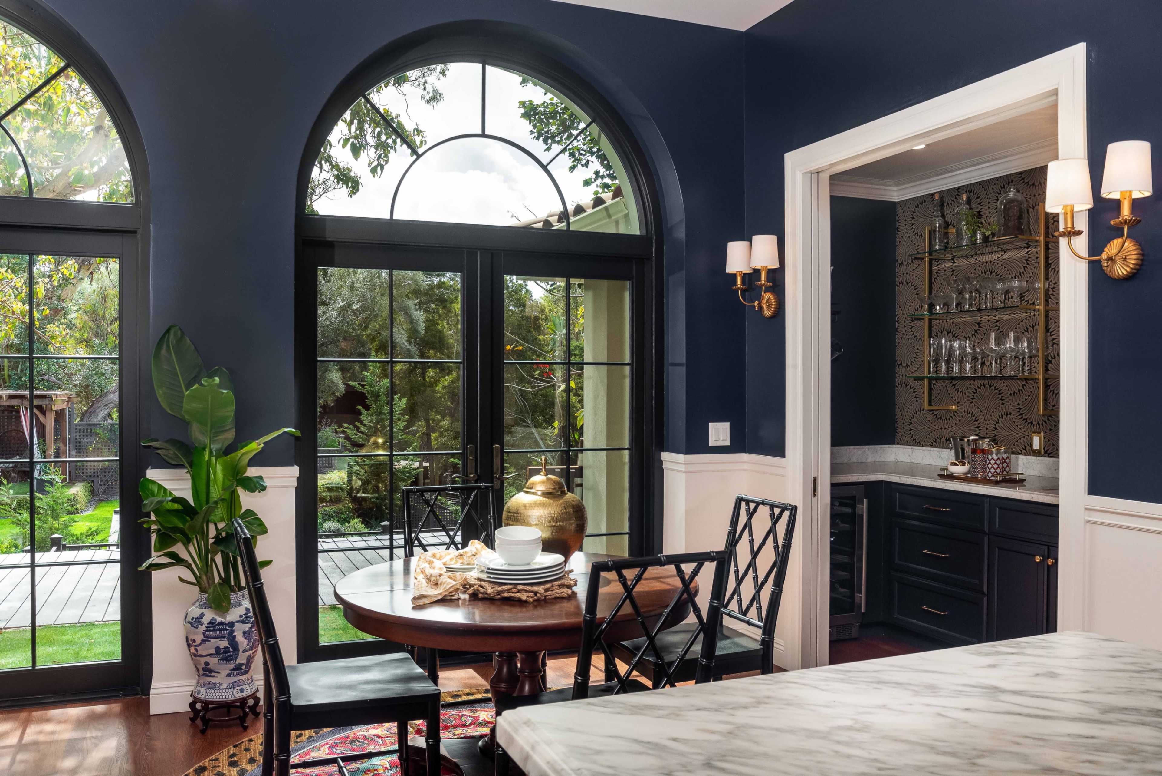 A dining area with a round table set for a meal, featuring large arched windows that overlook a lush garden.