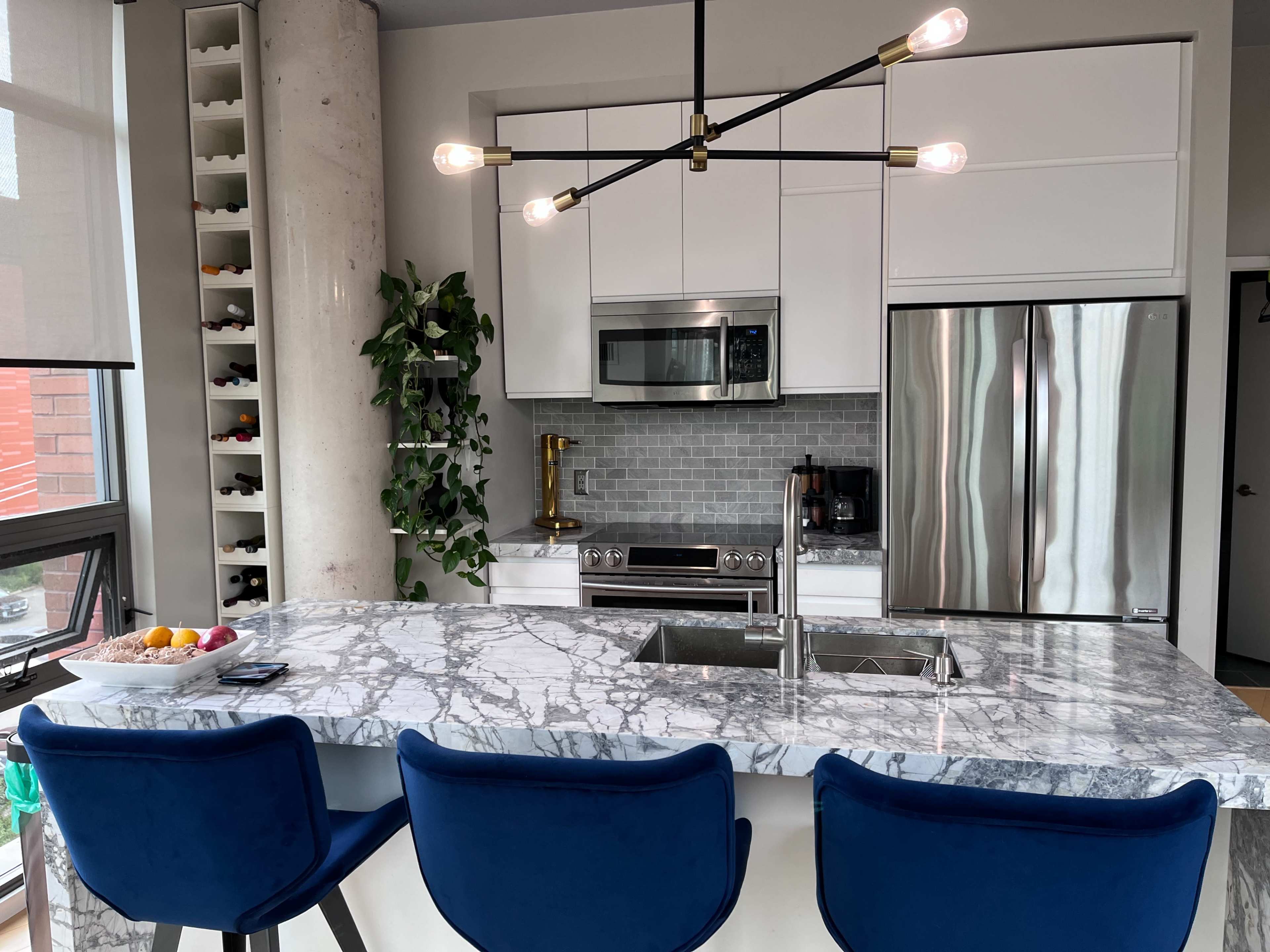 The image shows a modern kitchen featuring a marble countertop island with three blue bar stools, stainless steel appliances, and a sleek light fixture.