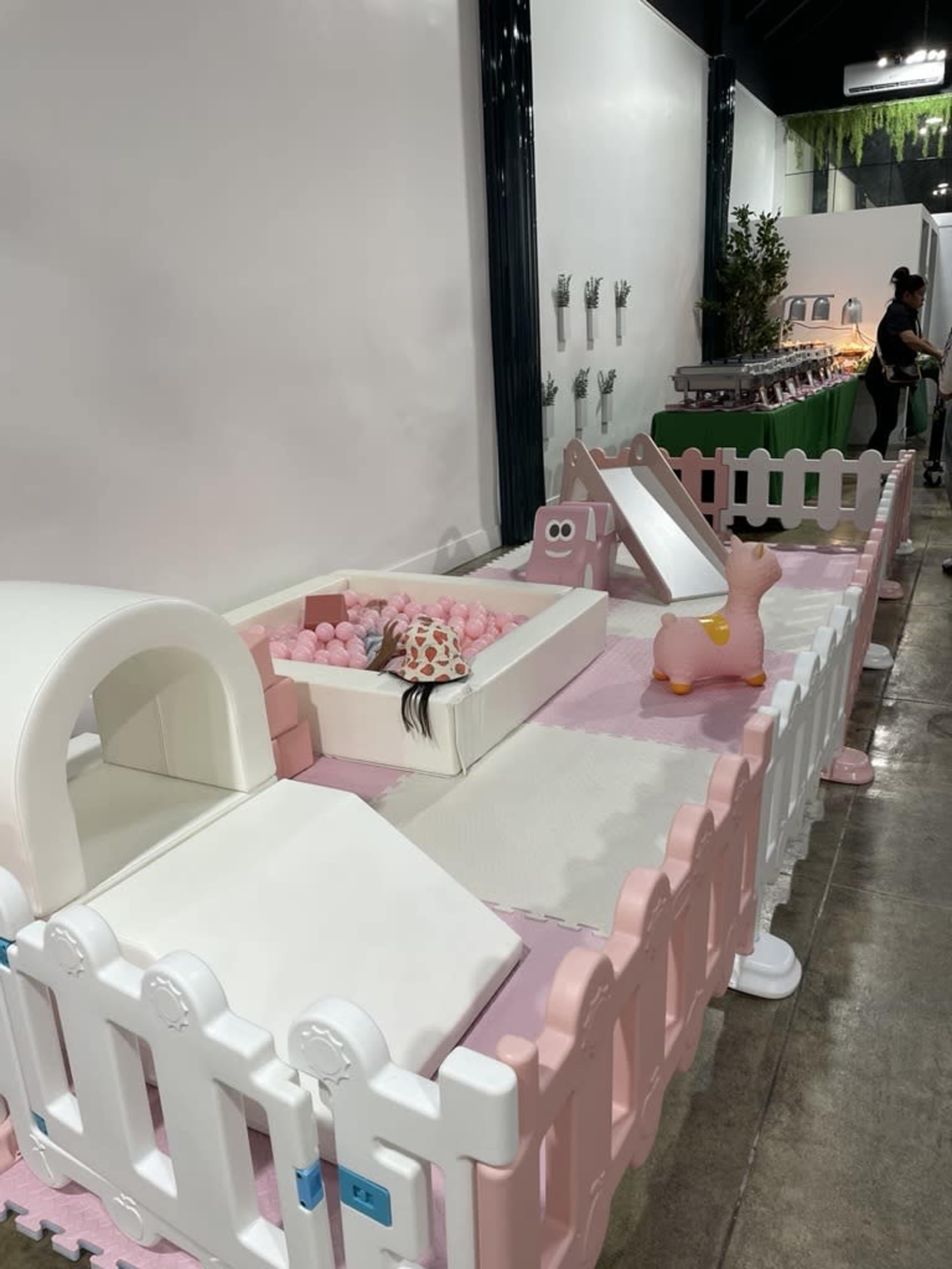 The image shows a play area enclosed by a white and pink plastic fence, featuring soft mats, a ball pit, and play equipment.