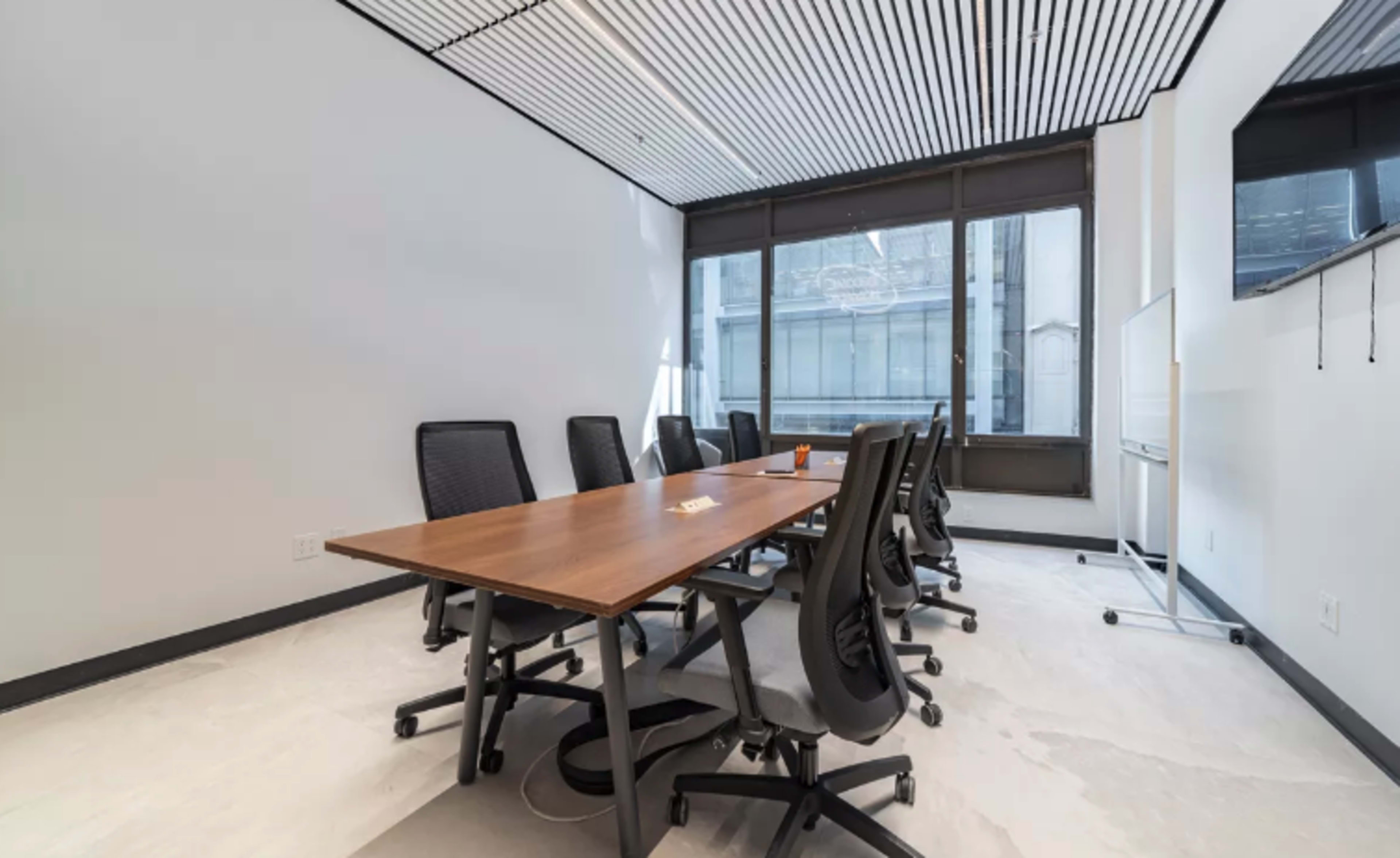 A modern conference room features a large wooden table surrounded by black ergonomic chairs, with large windows providing natural light.