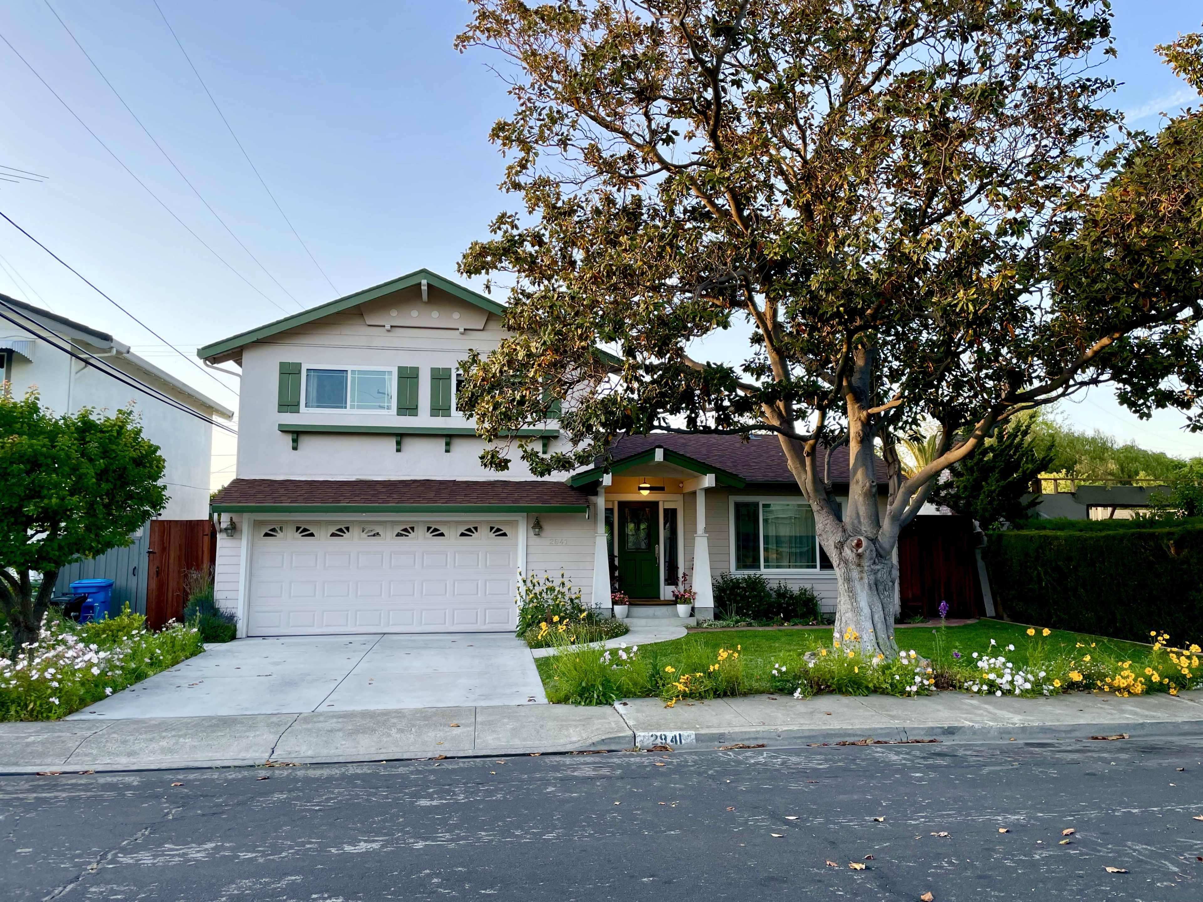 A two-story house with green shutters and a large tree in the front yard is situated on a quiet street.