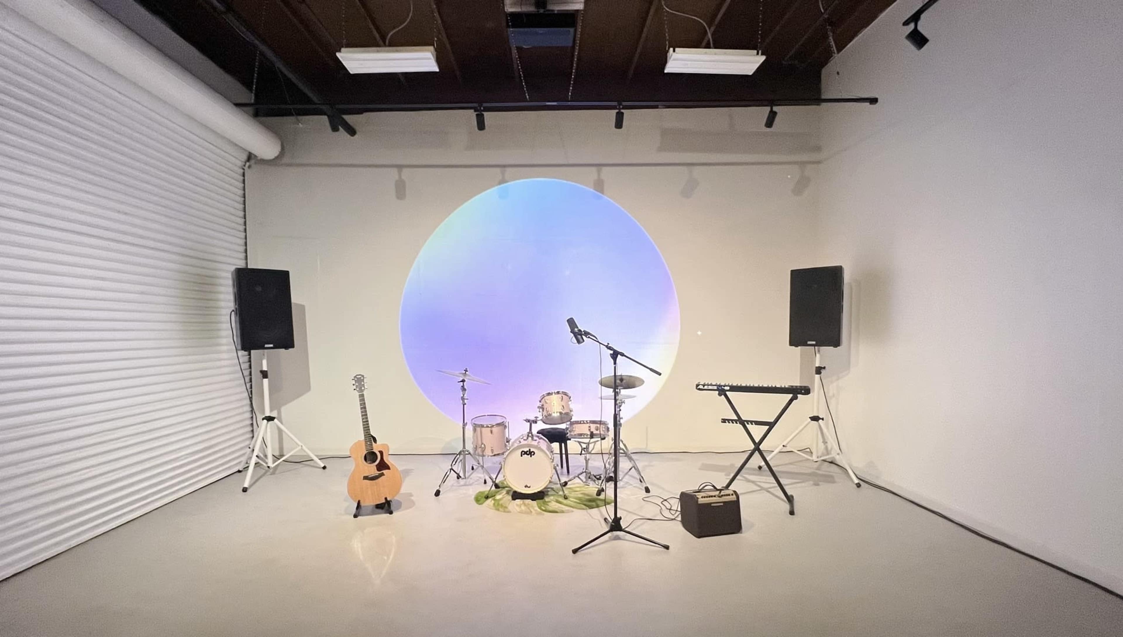 The image shows a minimalist performance space featuring a drum set, guitar, keyboard, and two speakers, with a large circular projection on the wall.