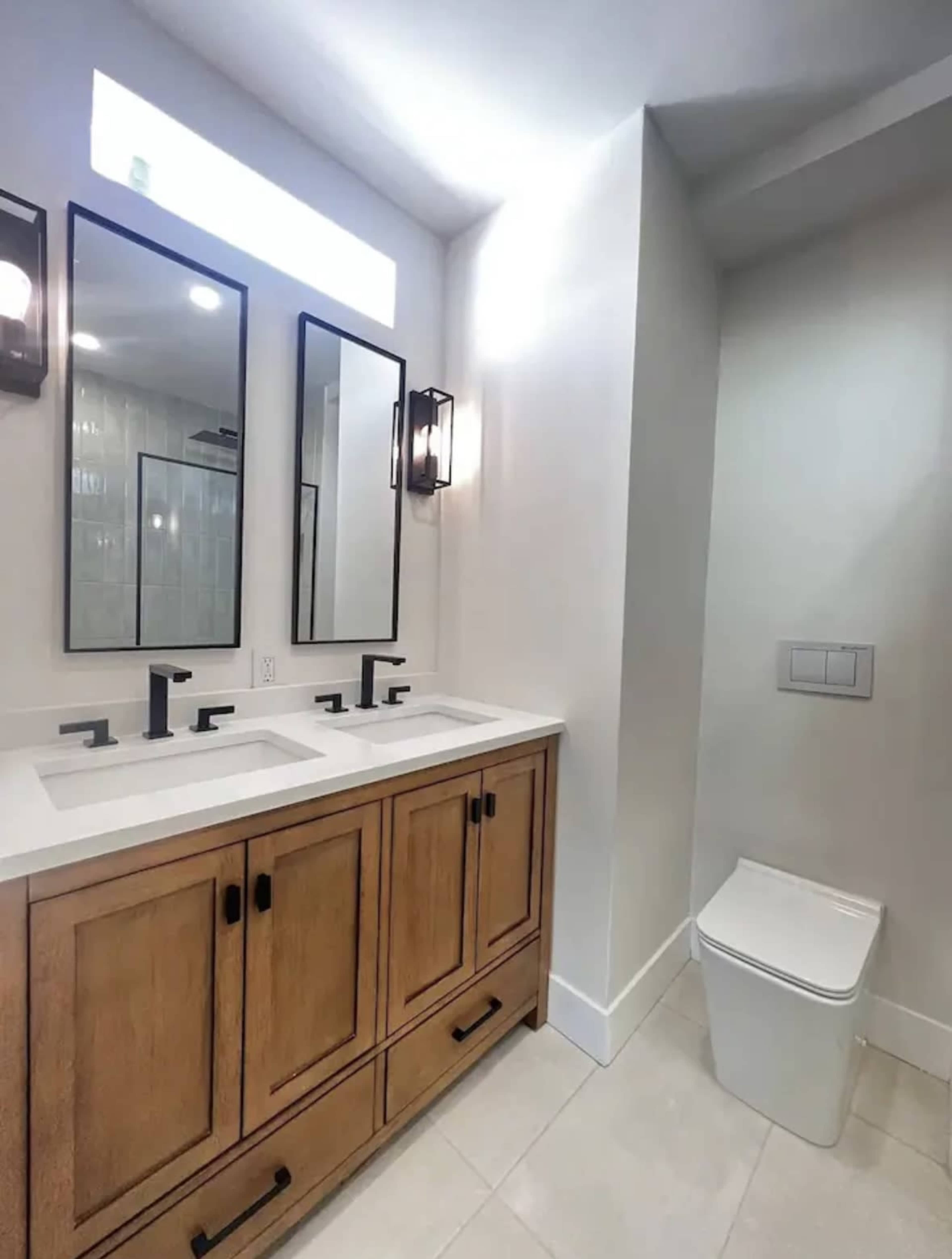 A modern bathroom featuring a double sink vanity with wooden cabinets, two mirrors, and a toilet in a separate corner.