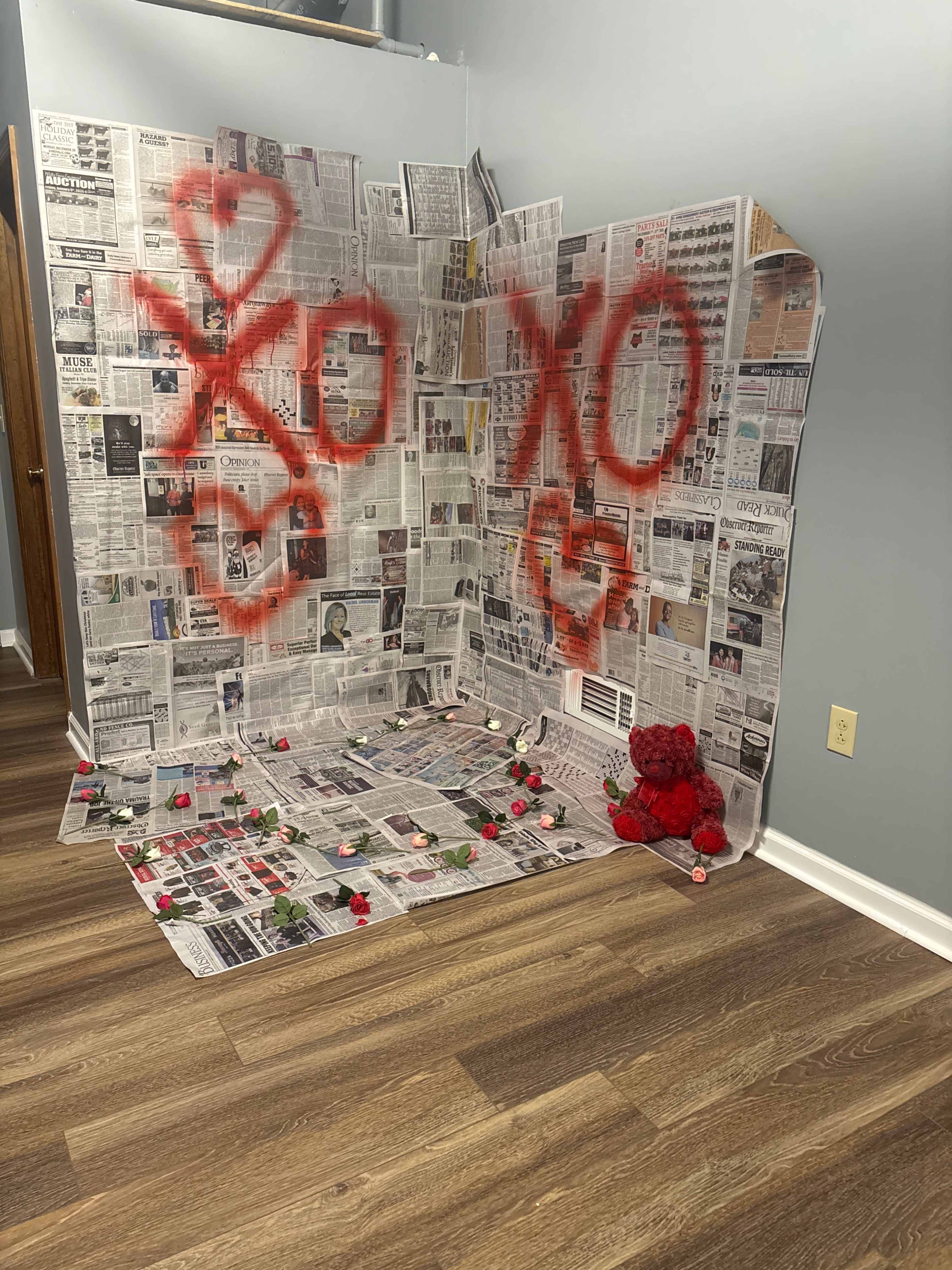 The image depicts a corner of a room with walls covered in newspaper, red spray-painted symbols, scattered rose petals on the floor, and a red stuffed bear in the foreground.