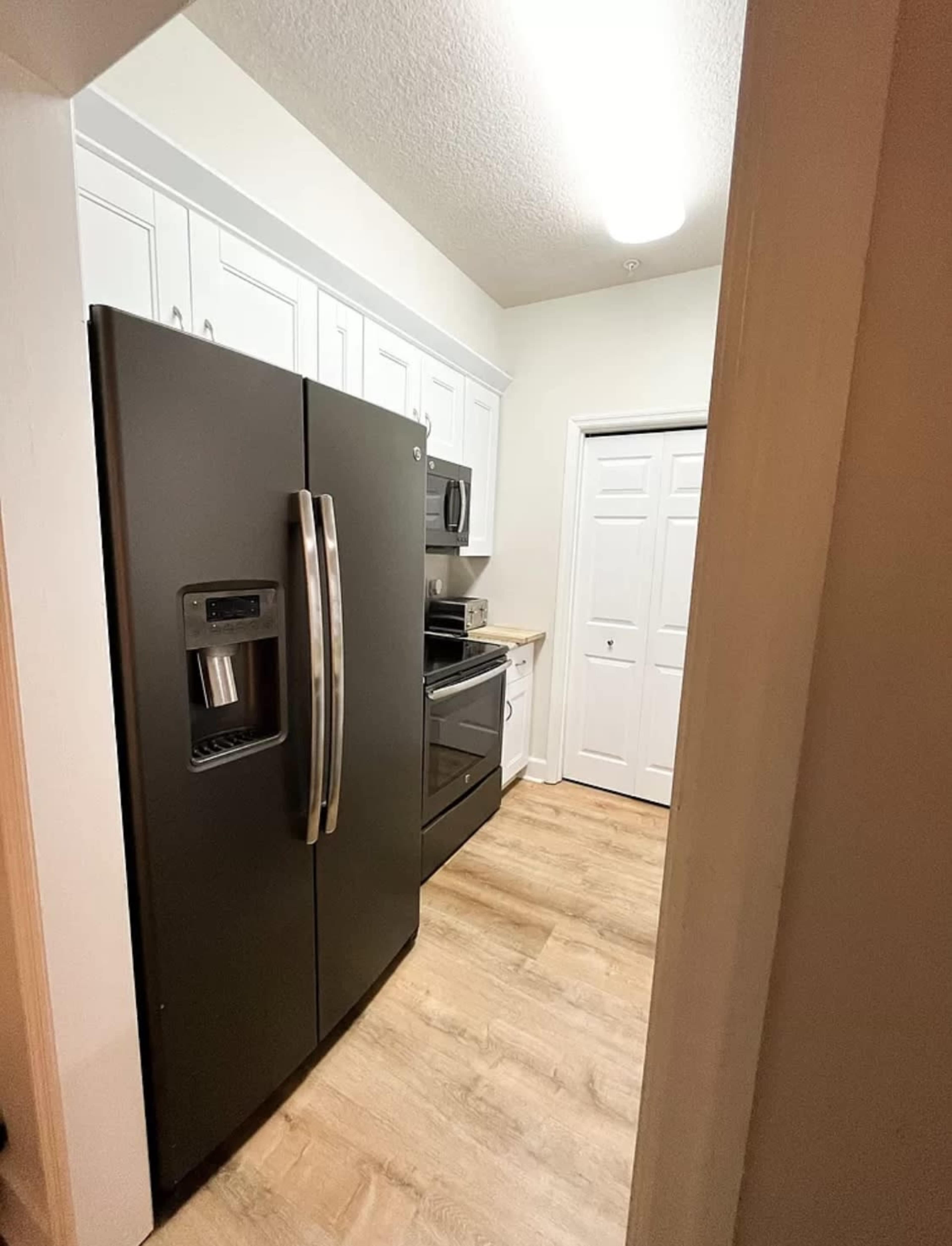 A black refrigerator and an oven are positioned in a narrow kitchen space with white cabinets and light wooden flooring.