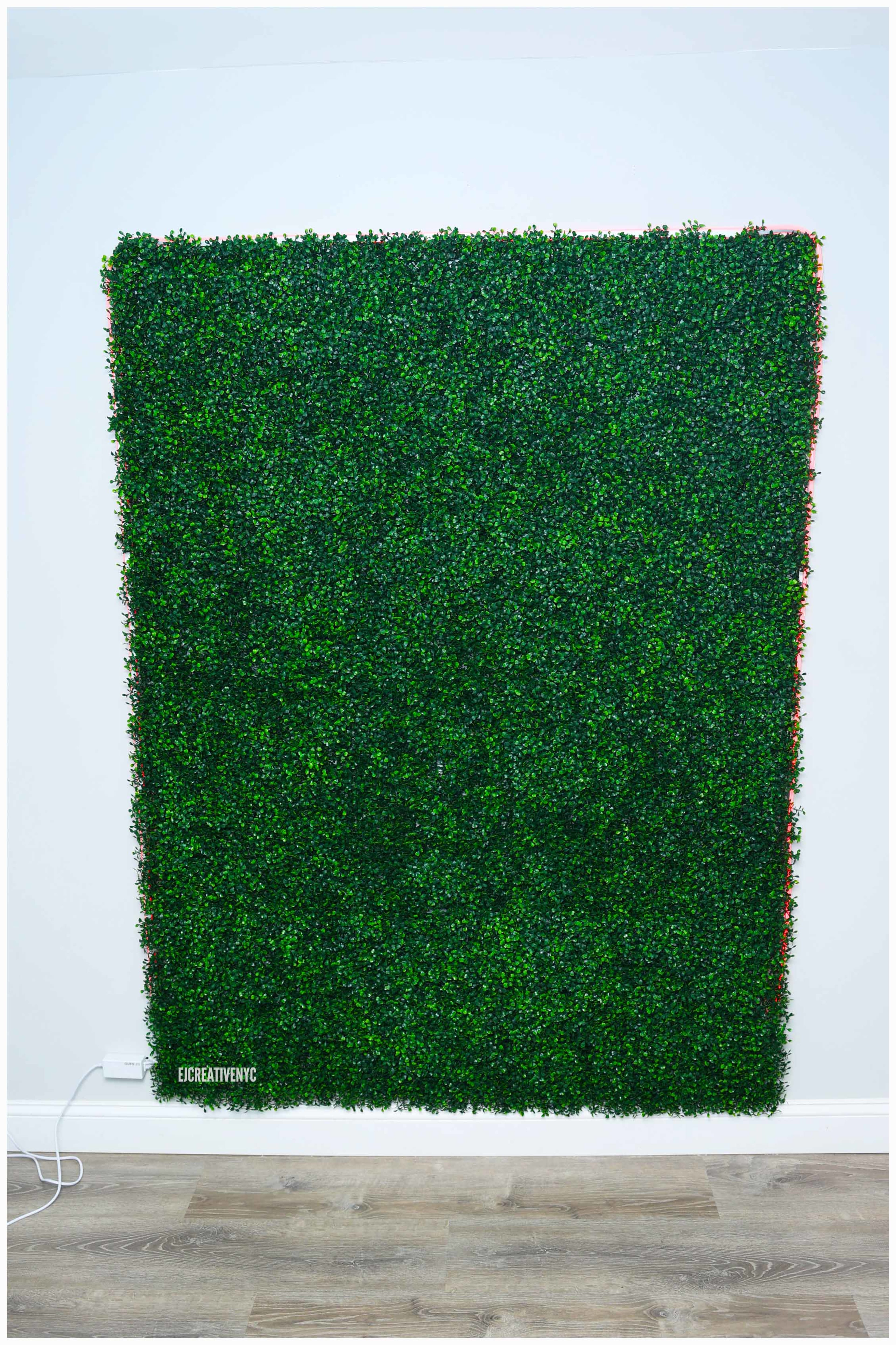 A rectangular panel of artificial greenery is mounted on a white wall above a wooden floor.