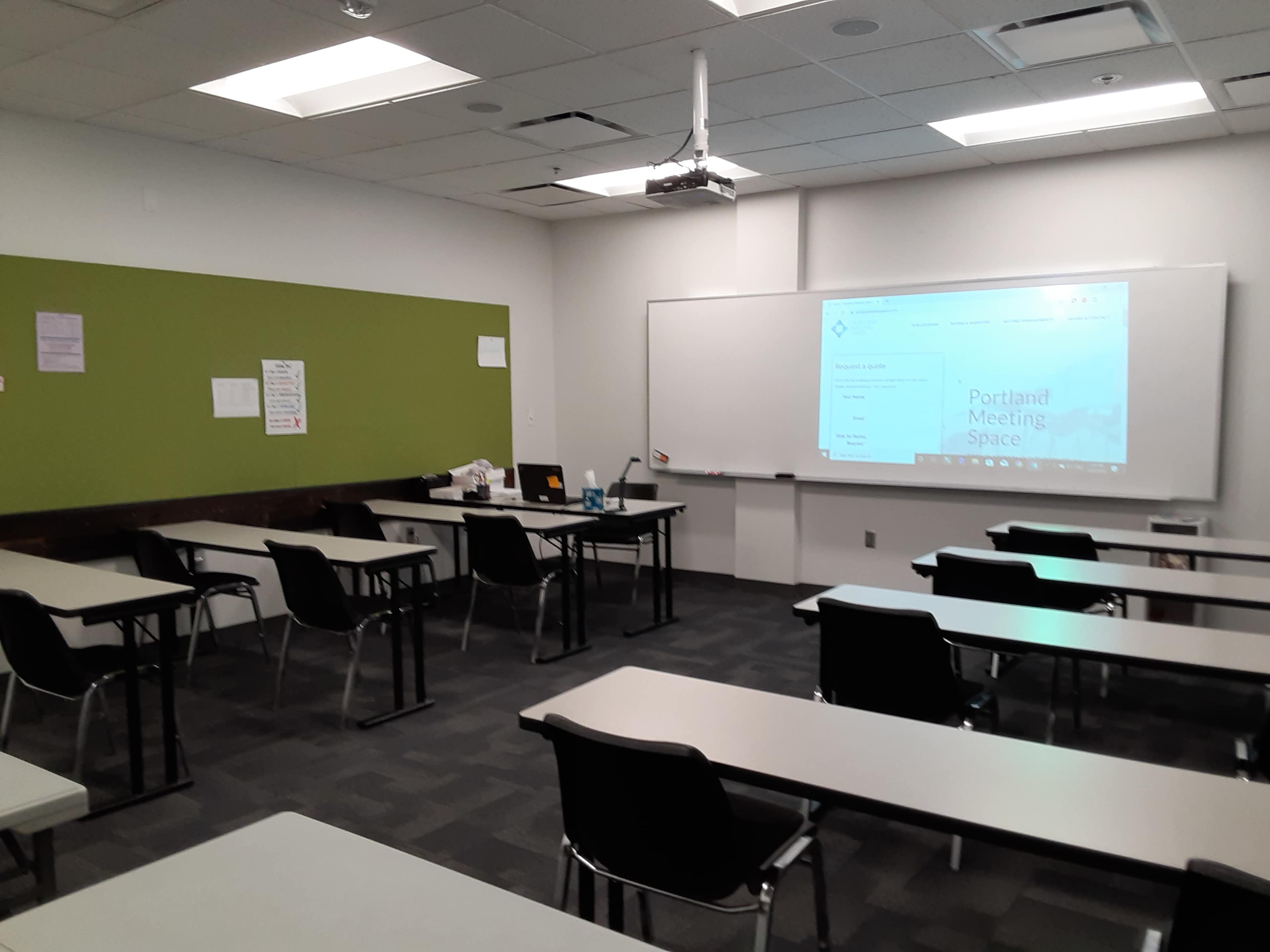A classroom features rows of desks facing a projected screen, with a green wall and minimal decorations.