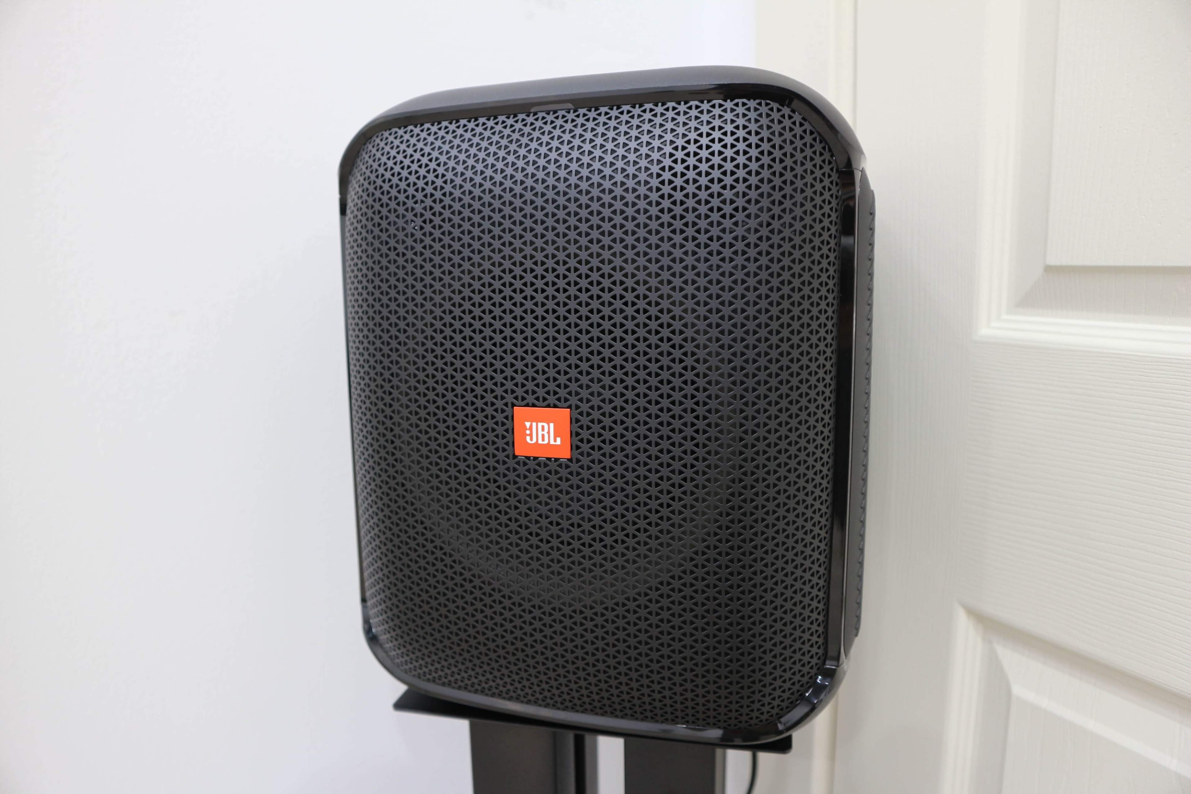A JBL speaker mounted on a wall, featuring a black exterior with a perforated grille and a prominent red logo.