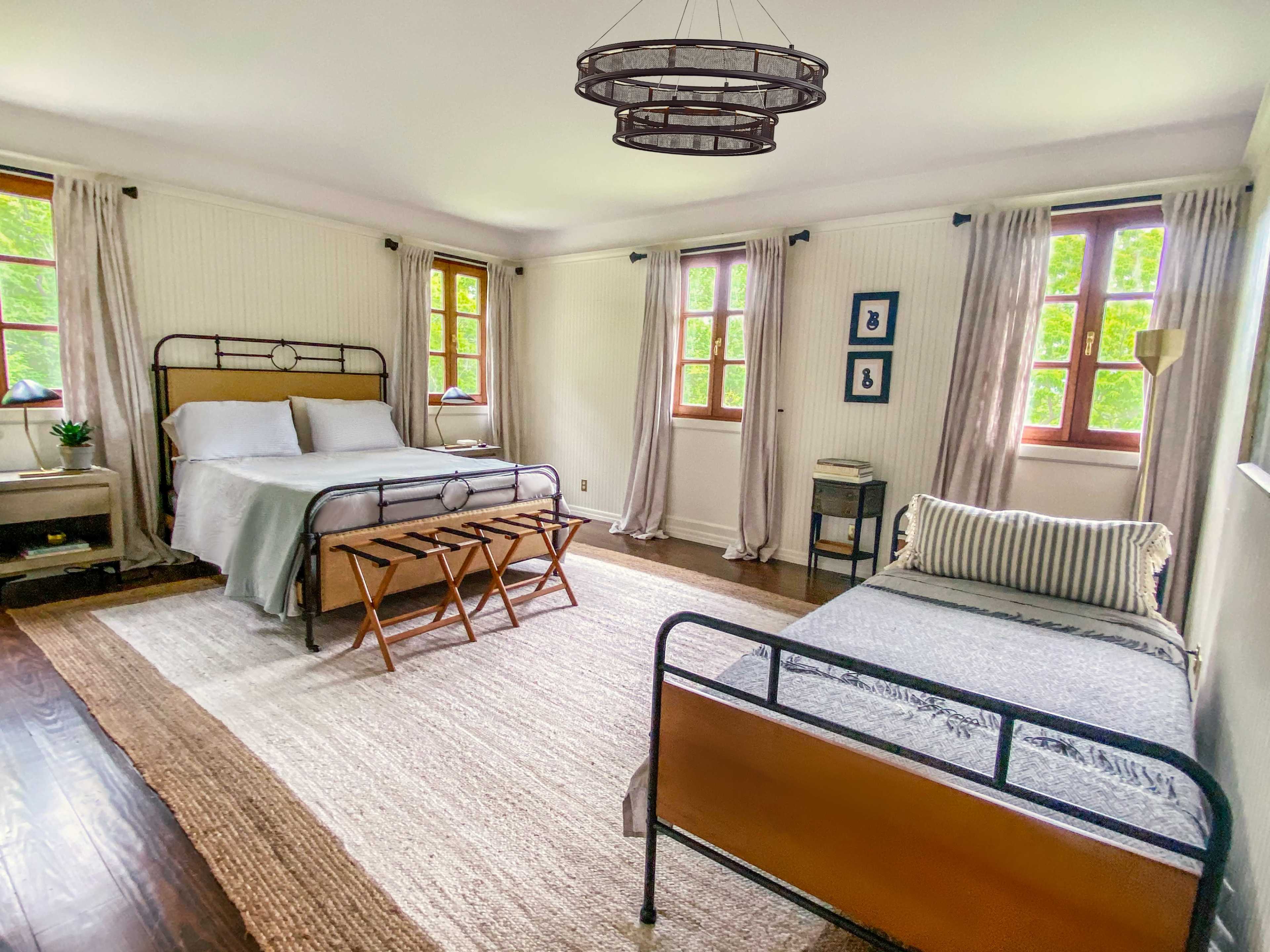 A spacious bedroom featuring a queen-sized bed, a twin bed, large windows with light drapes, and wooden flooring.