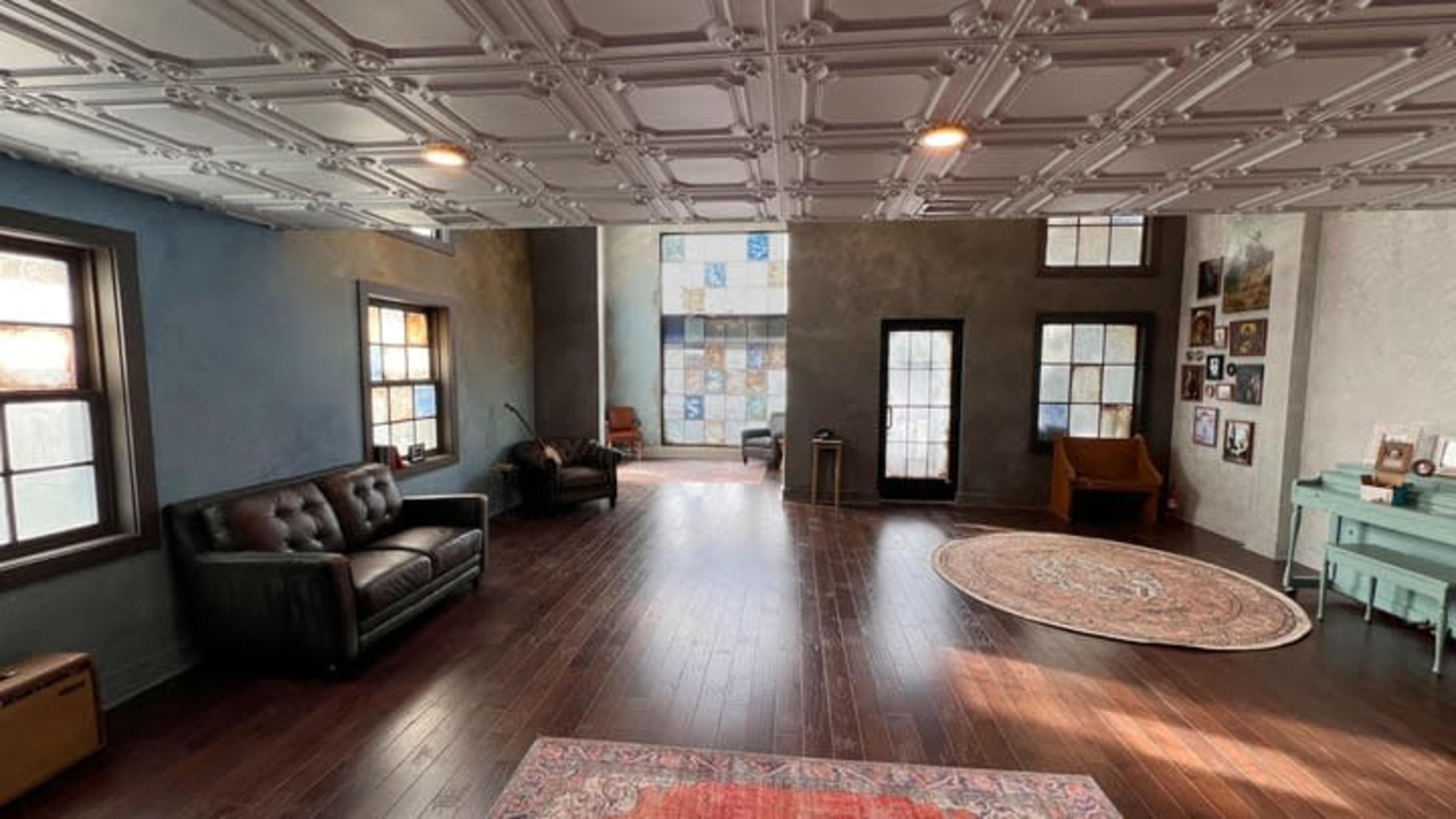 The image shows a spacious interior room with wooden floors, multiple windows, a variety of seating options, and decorative elements on the walls.