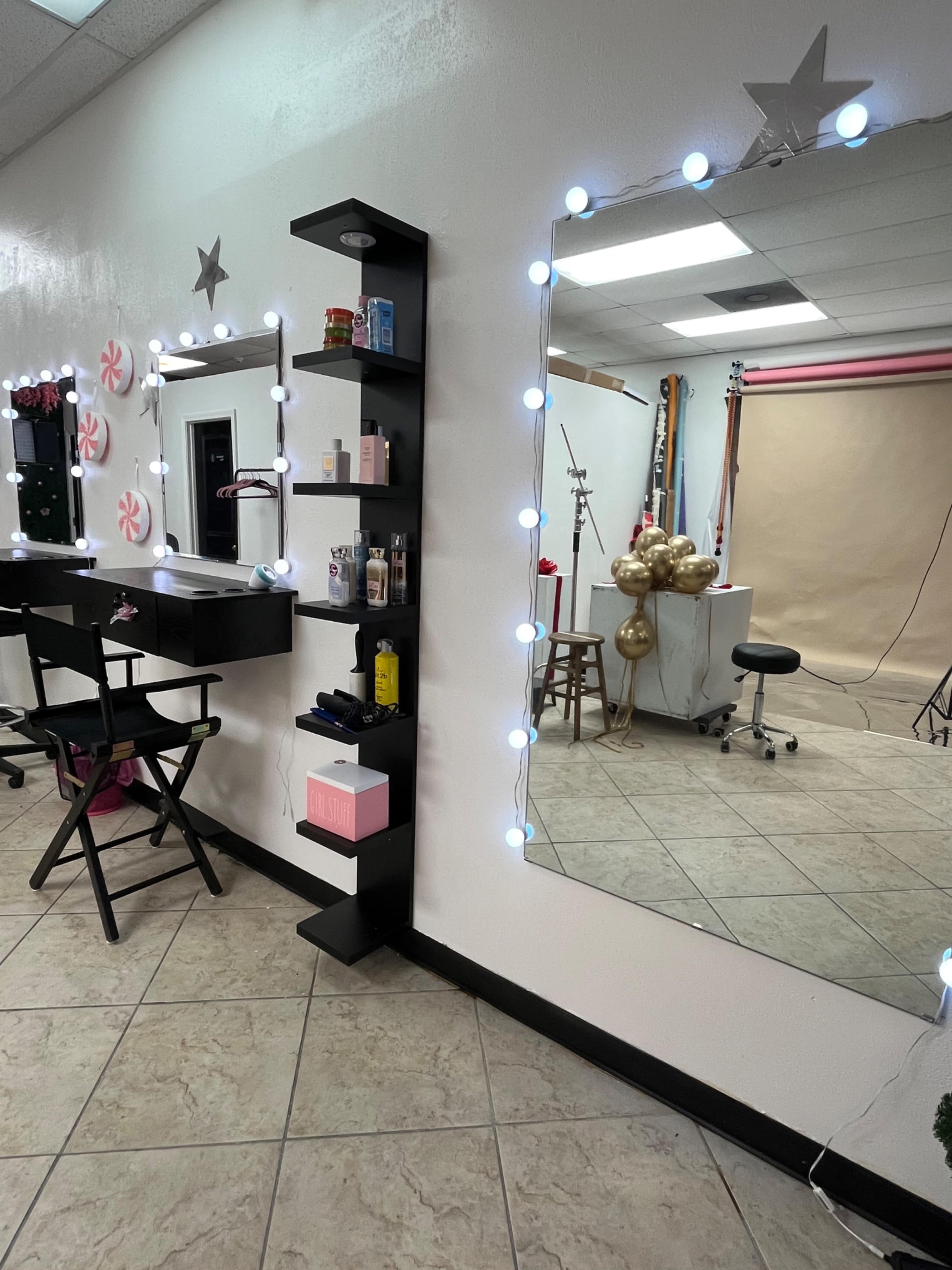 The image shows a well-lit beauty salon with a vanity area featuring a mirror surrounded by lights, shelves with beauty products, and a photography backdrop in the background.