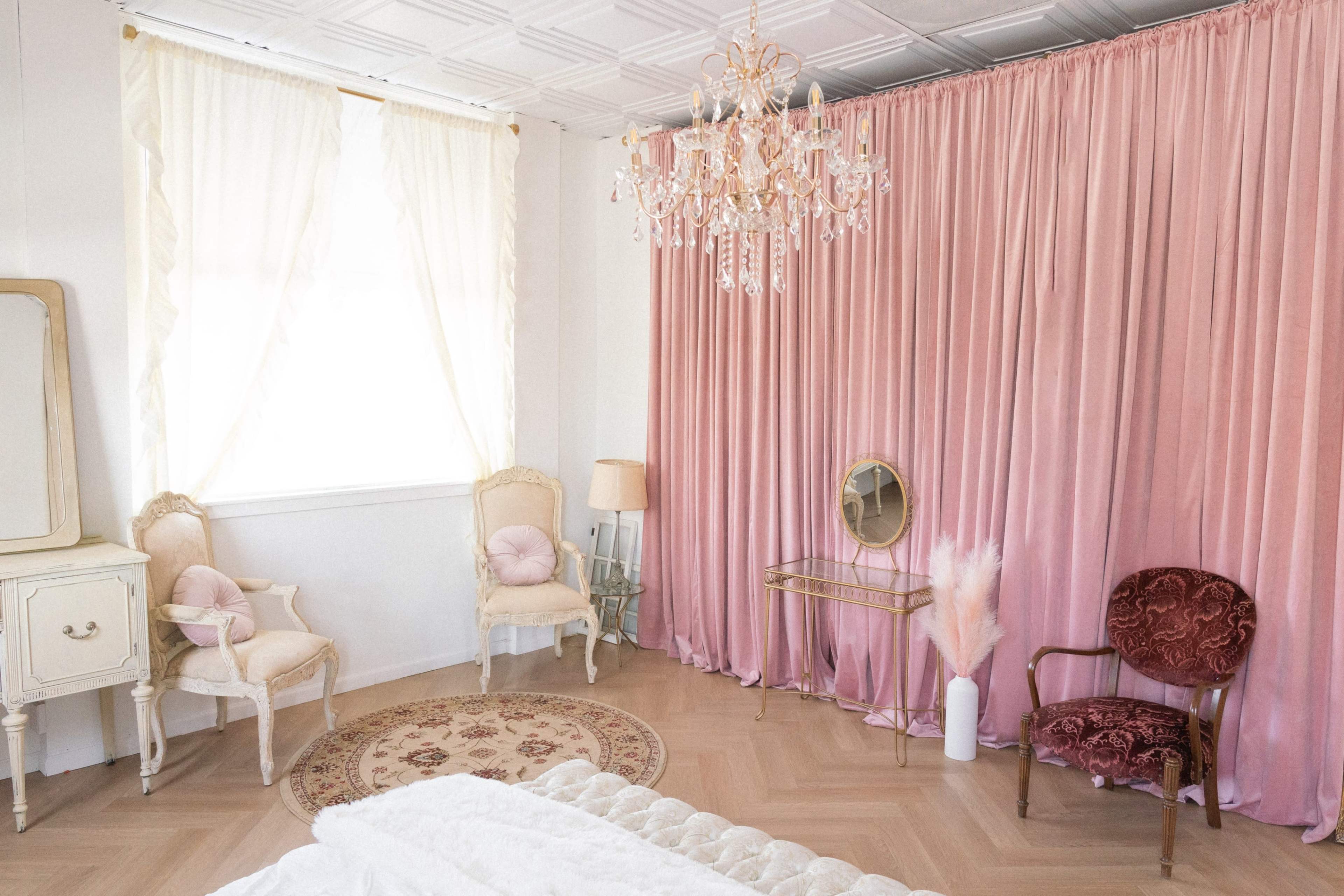 The image shows a softly lit room featuring a pink velvet curtain backdrop, a chandelier, a round patterned rug, and vintage furniture including a mirror and two chairs.