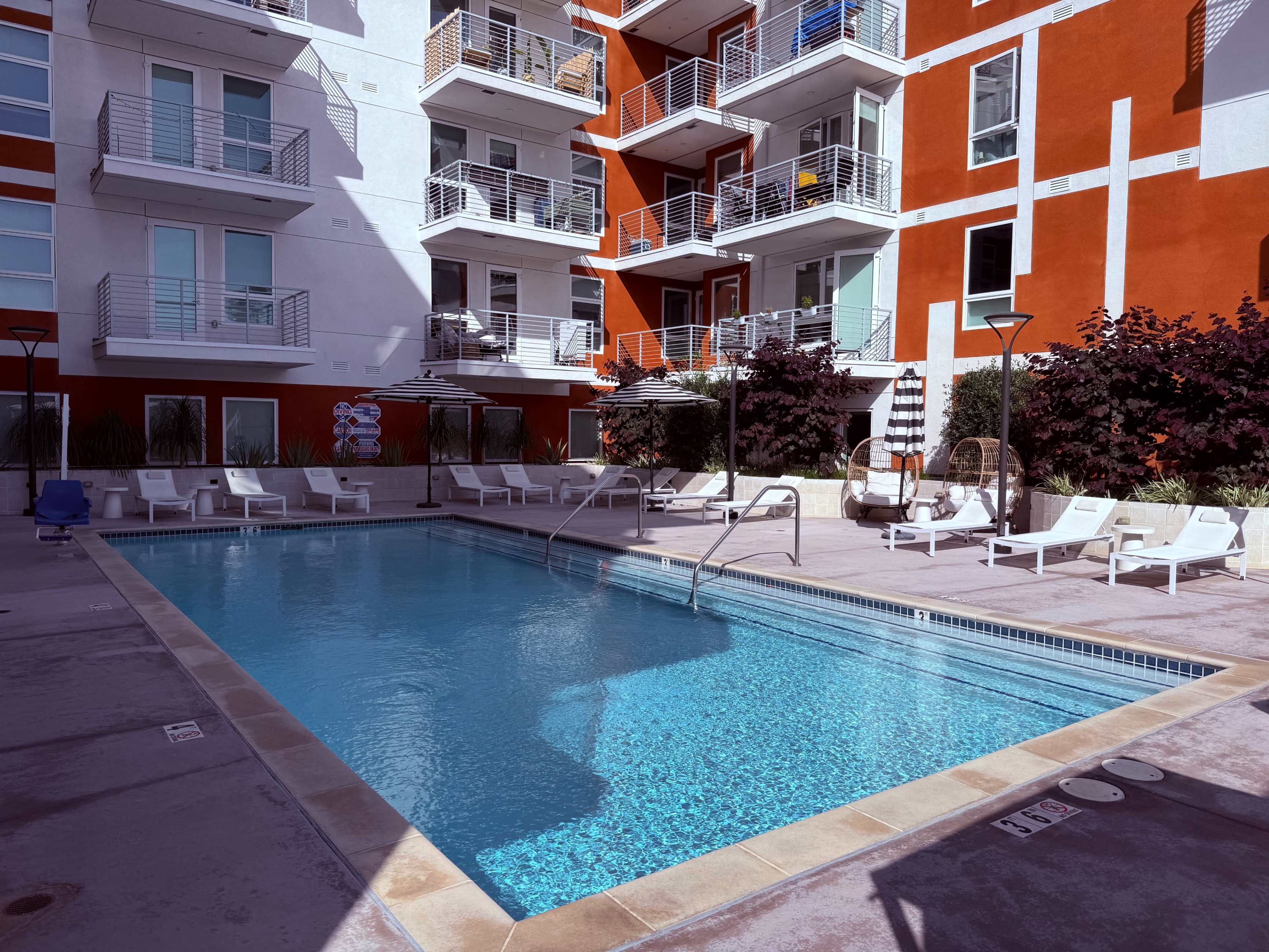 The image shows a rectangular swimming pool surrounded by lounge chairs and balconies of a residential building with a red and white exterior.
